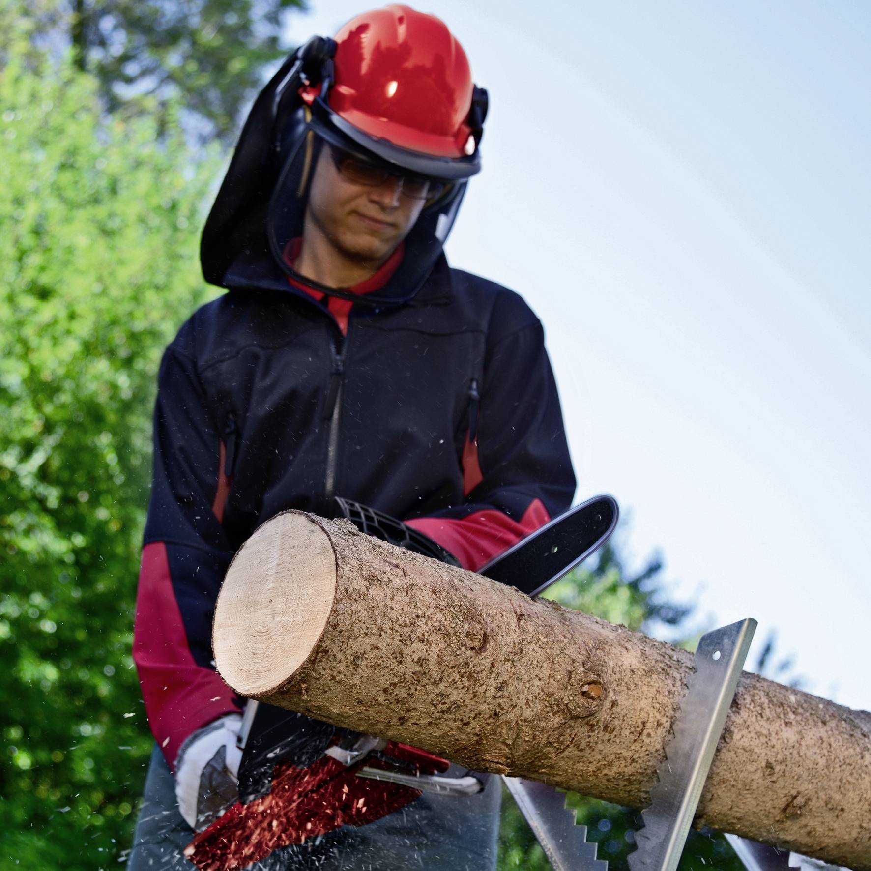 Mężczyzna w odzieży ochronnej piłuje piłą mechaniczną pień drzewa na wolnym powietrzu. W tle widoczne są drzewa i błękitne niebo.