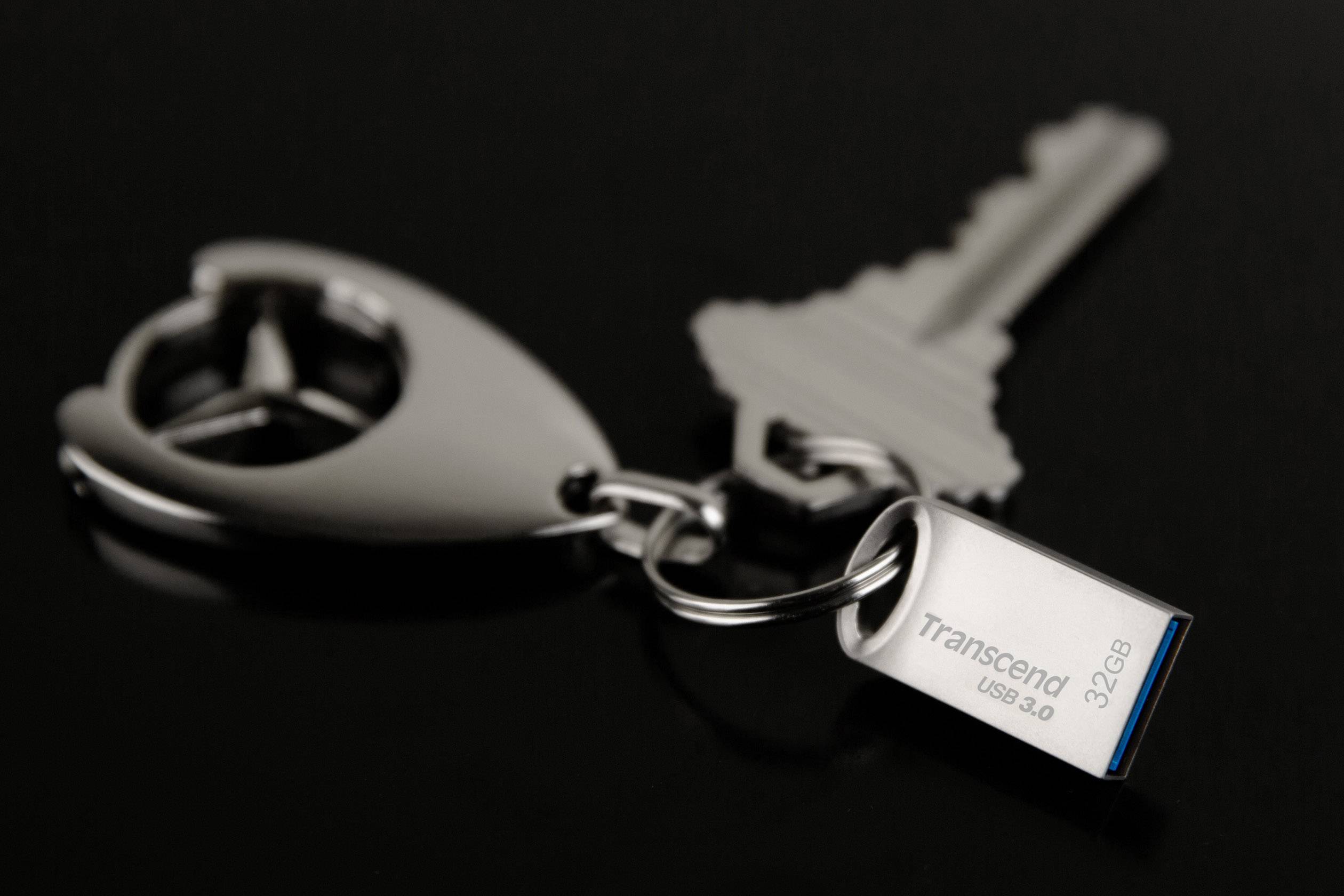 Brelok z logo Mercedes oraz pendrive'em Transcend o pojemności 32 GB, w standardzie USB 3.0, sfotografowany na czarnym tle.