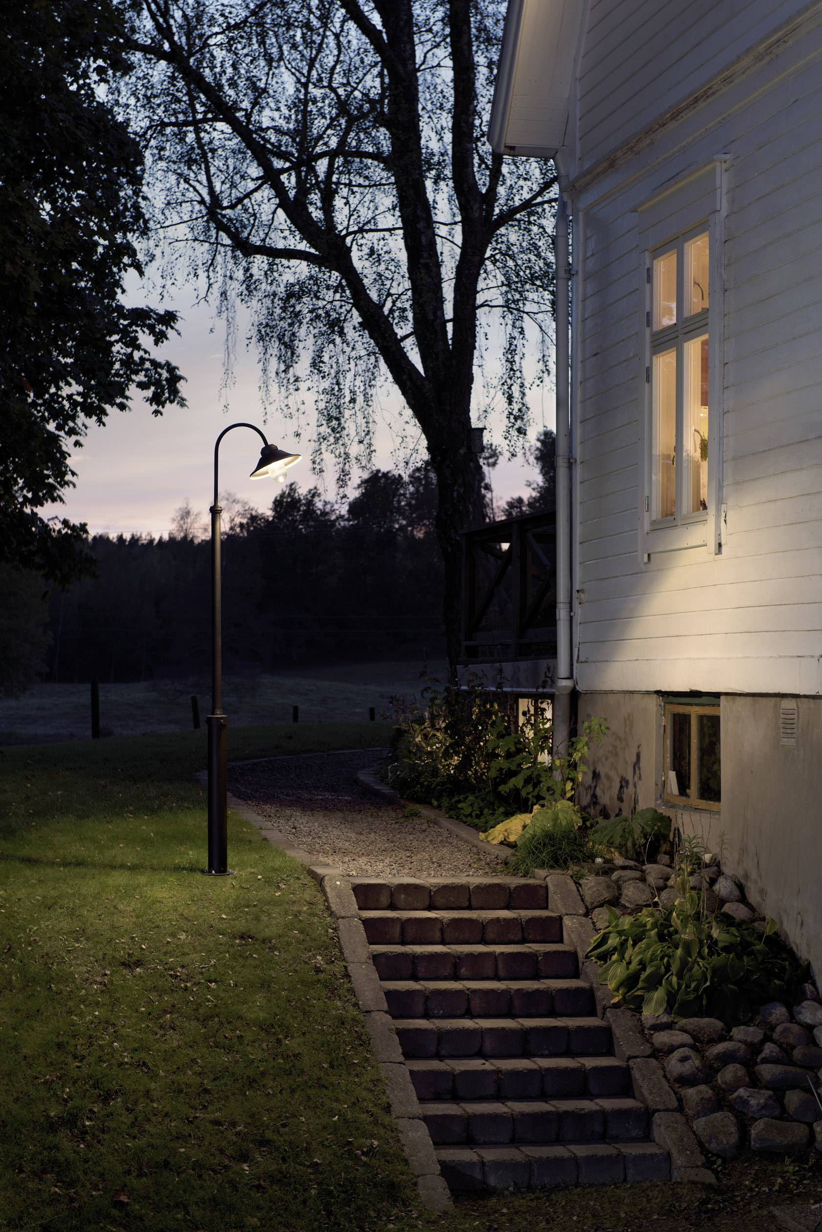 Biały dom o zmierzchu z oświetlonym oknem i latarnią, przed schodami w ogrodzie, otoczony drzewami i trawnikiem.