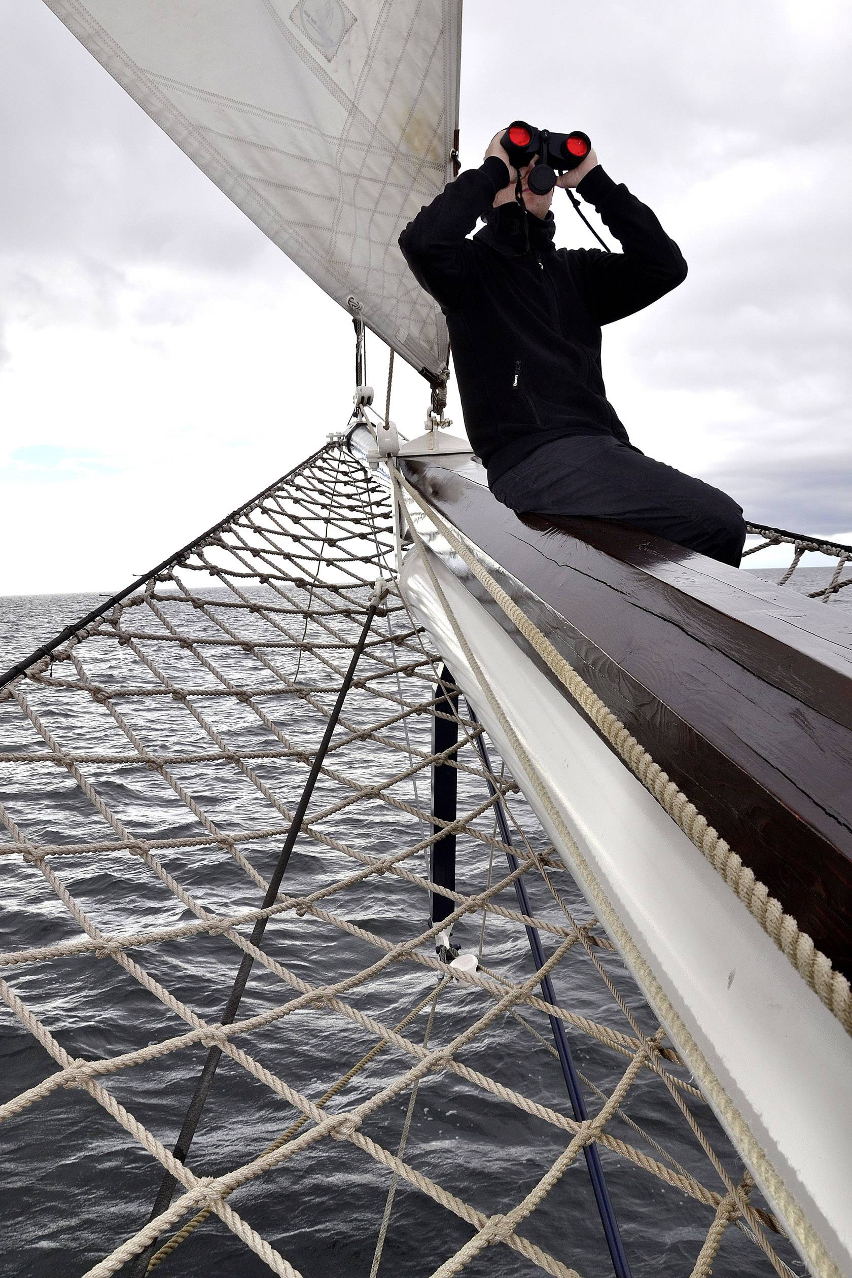 Osoba siedzi na dziobie żaglówki i patrzy przez lornetkę na morze. Niebo jest zachmurzone, a morze spokojne.