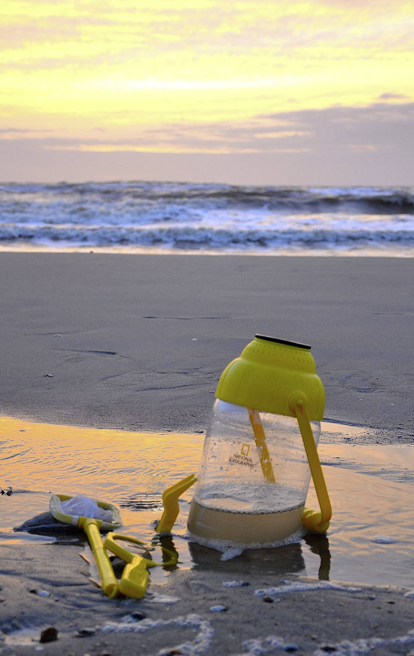 Żółty plastikowy pojemnik i zabawka leżą na piasku na plaży. W tle widać morze o zachodzie słońca.