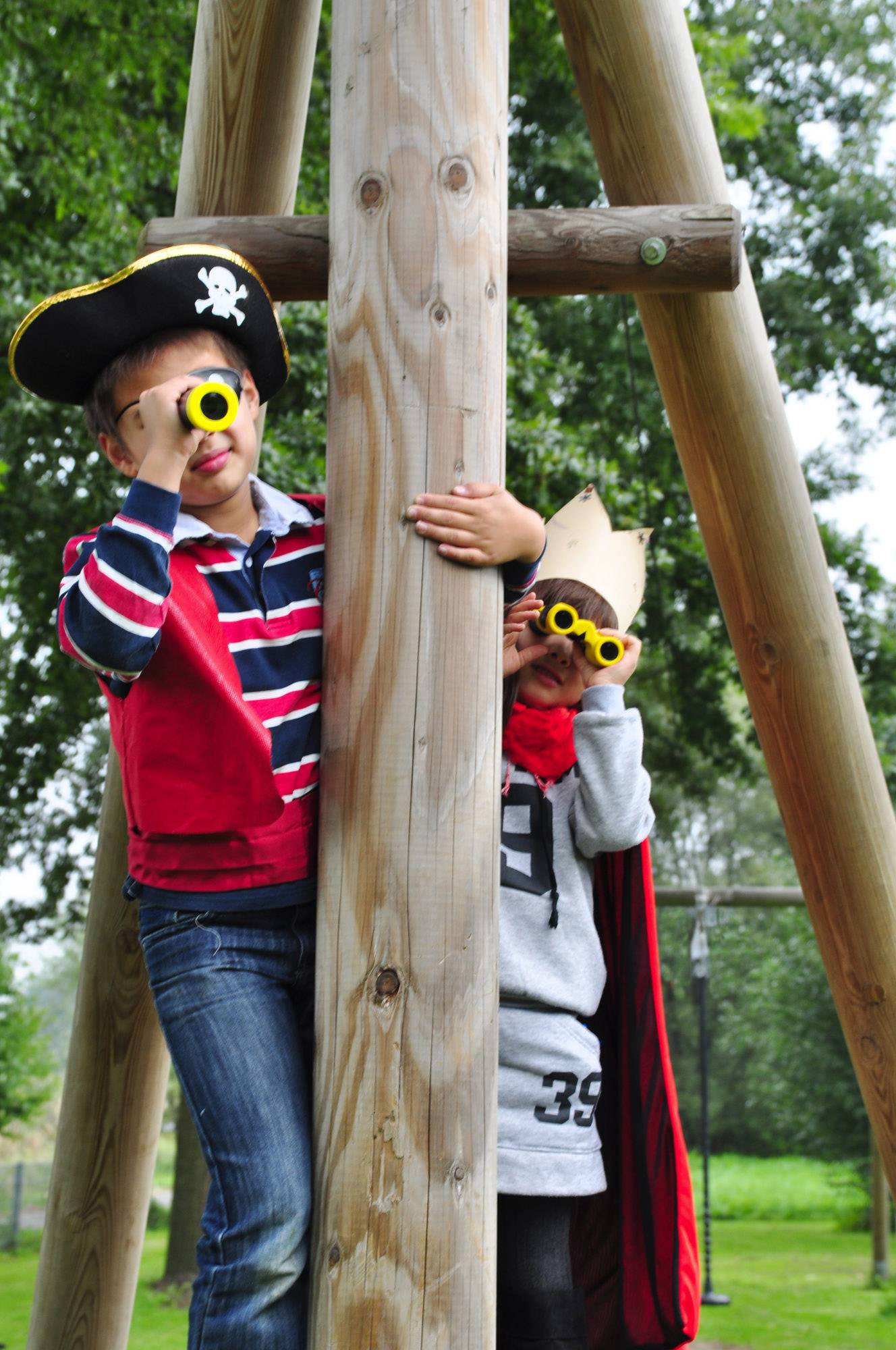 Dwoje dzieci, przebrane za pirata i króla, bawi się na drewnianym rusztowaniu na dworze. Spoglądają przez zabawkowe lornetki.