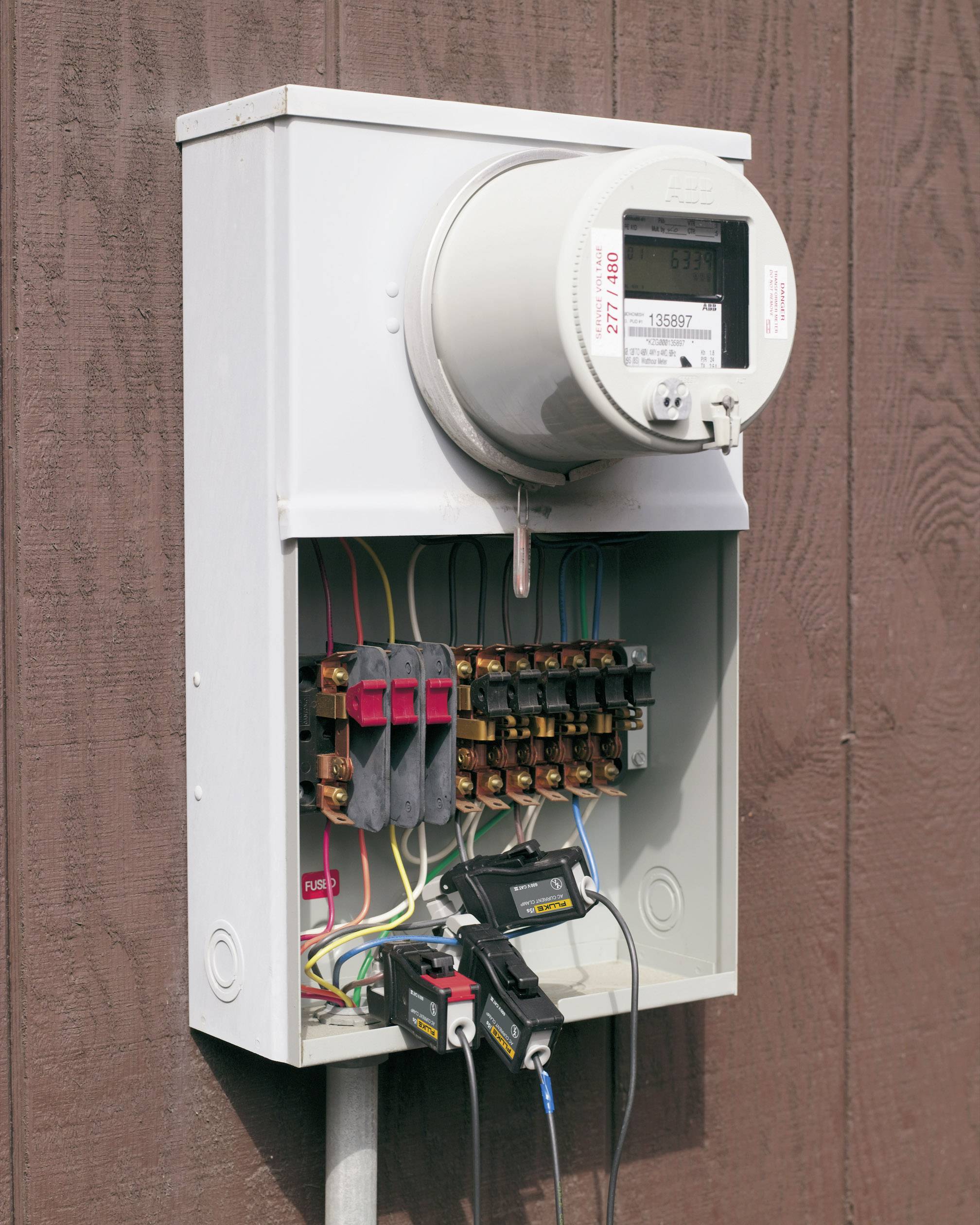 Elektryczna skrzynka licznikowa na drewnianej ścianie, z widocznymi kolorowymi przewodami i bezpiecznikami wewnątrz obudowy.