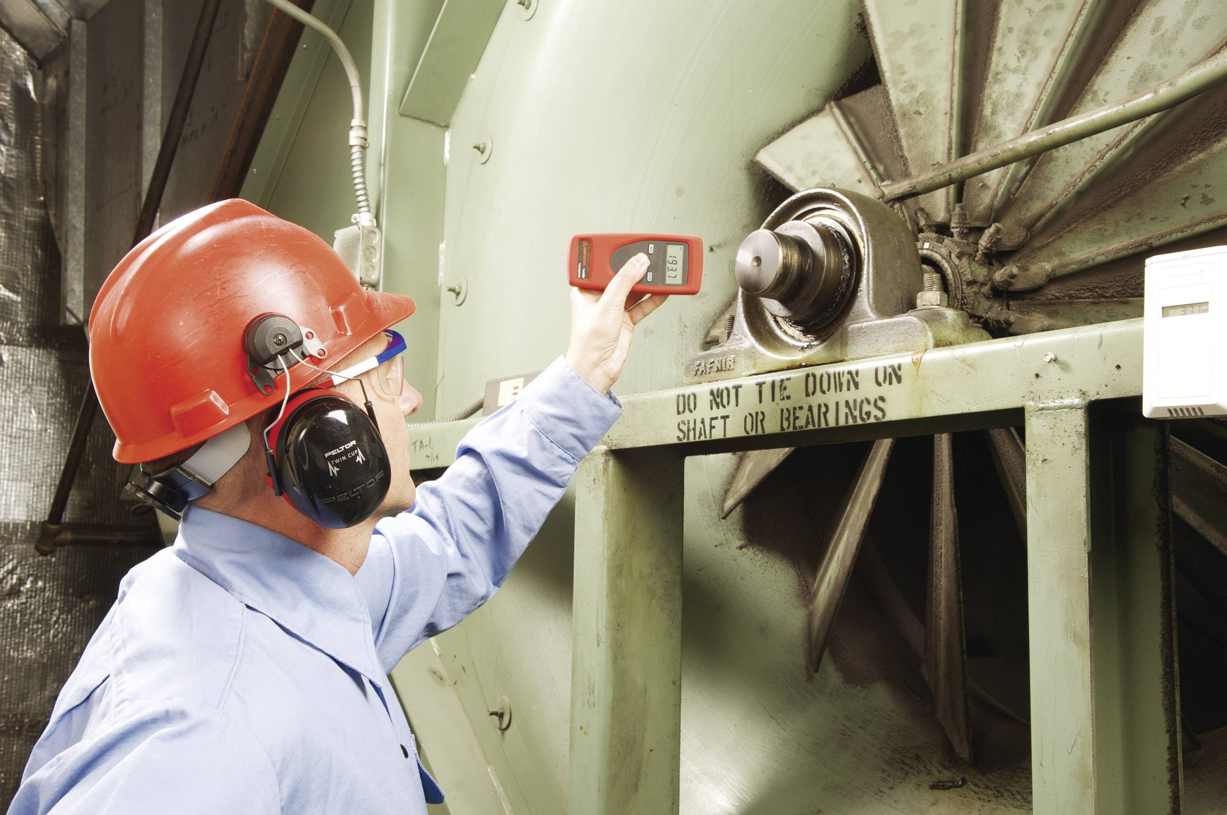 Pracownik w kasku i ochronnikach słuchu sprawdza urządzeniem wentylator przemysłowy. Widoczne są środki bezpieczeństwa.