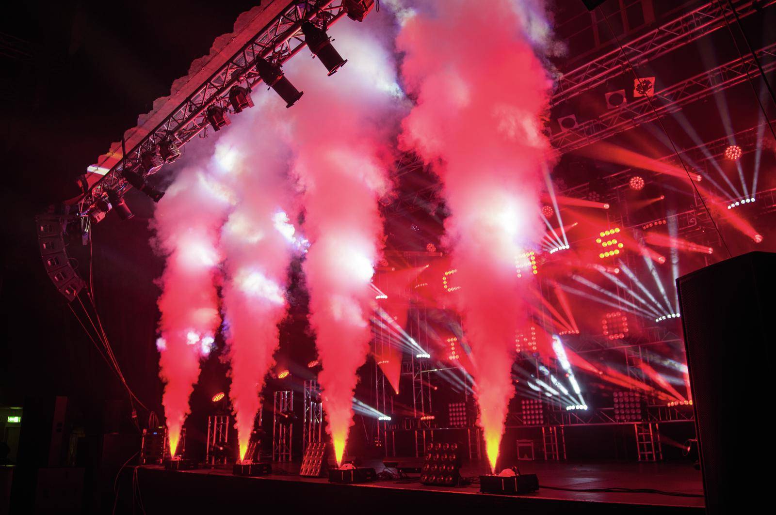 Scena z czerwonymi efektami świetlnymi i maszynami dymnymi, które wyrzucają dym w powietrze. Oświetlenie tworzy dramatyczną atmosferę.