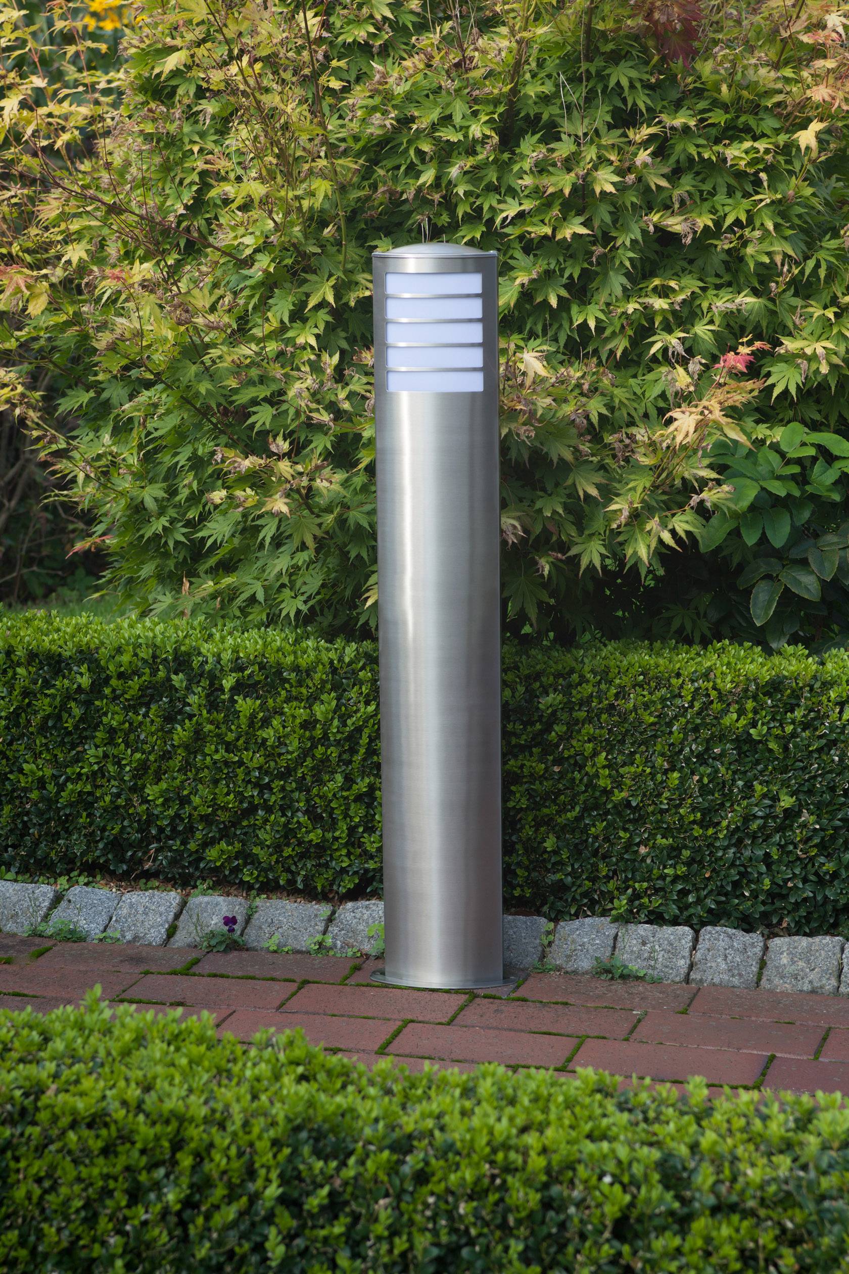 Nowoczesna lampa ogrodowa z metalu stoi w starannie utrzymanym ogrodzie, a w tle widoczne są rośliny i żywopłoty.