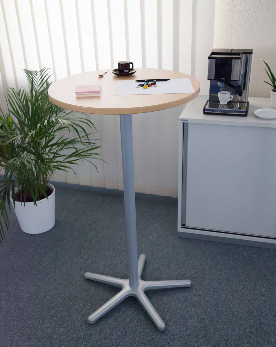 Stolik do stania w biurze z filiżanką kawy, notatnikiem i długopisami. W tle ekspres do kawy i rośliny.