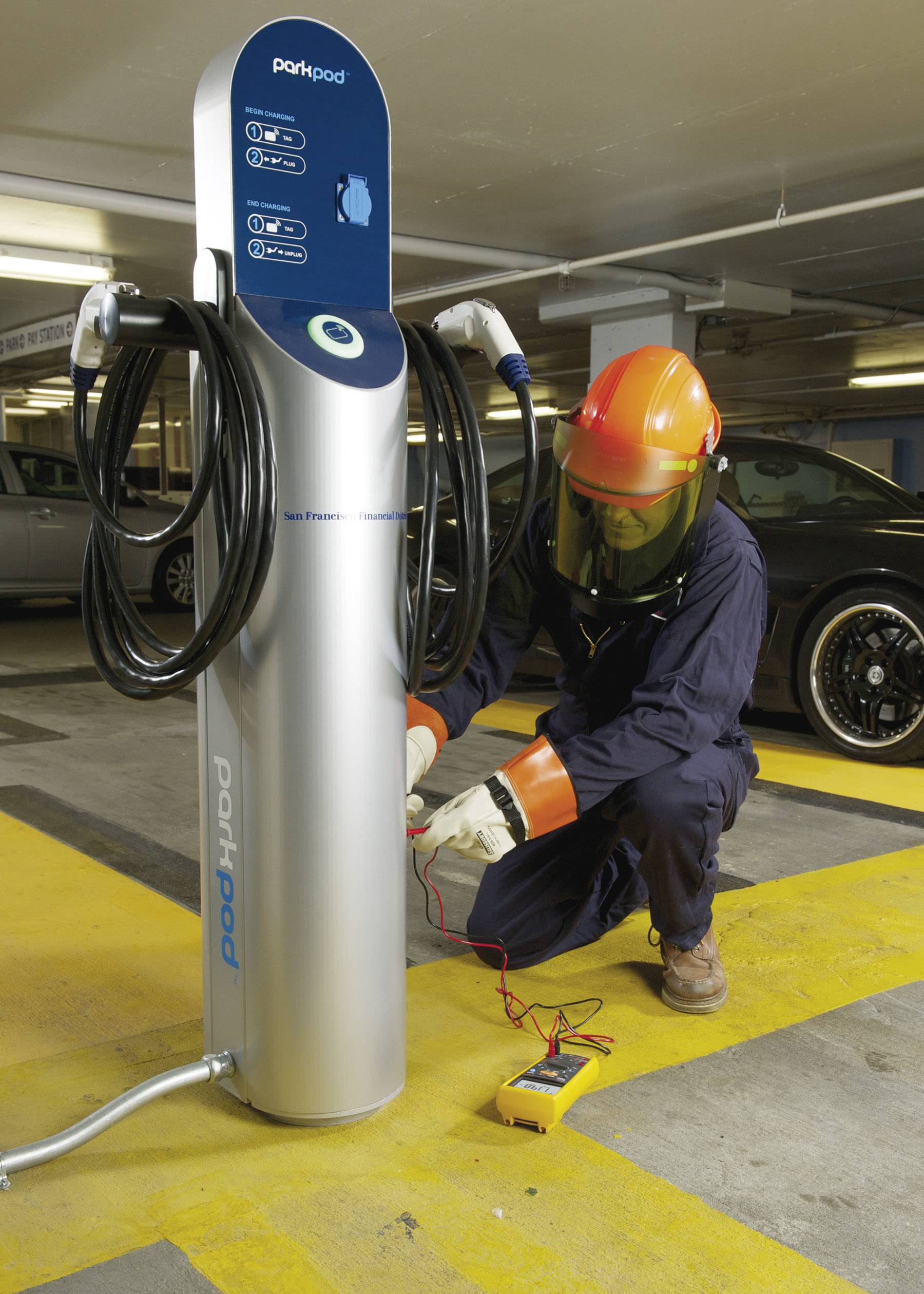 Technik w odzieży ochronnej sprawdza stację ładowania samochodu elektrycznego w podziemnym parkingu. Na podłodze leży przyrząd pomiarowy.