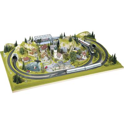 Miniaturowa krajobraz z trasą kolejową, dwoma pociągami, wieloma budynkami i drzewami, małymi wzgórzami oraz jeziorem z żaglówką.
