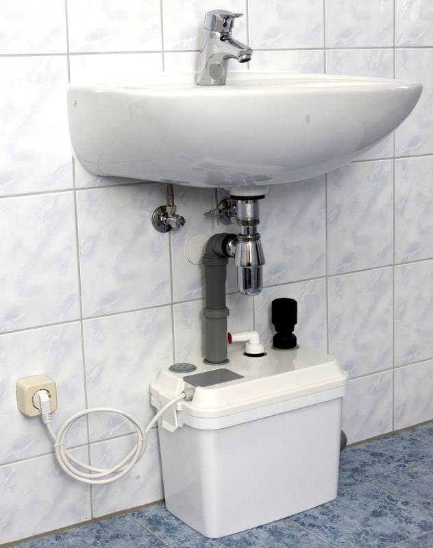 Umywalka, pod nią mała pompka do ścieków i widoczne rury, na tle wyłożonej kafelkami ściany. Po lewej stronie gniazdo elektryczne.