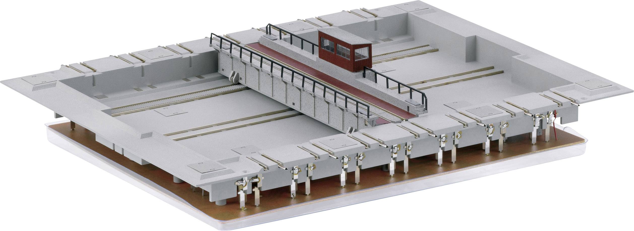 Szara miniaturowa makieta platformy kolejowej z przęsłami, z czerwonym budynkiem kontrolnym pośrodku i szczegółowymi elementami po bokach.