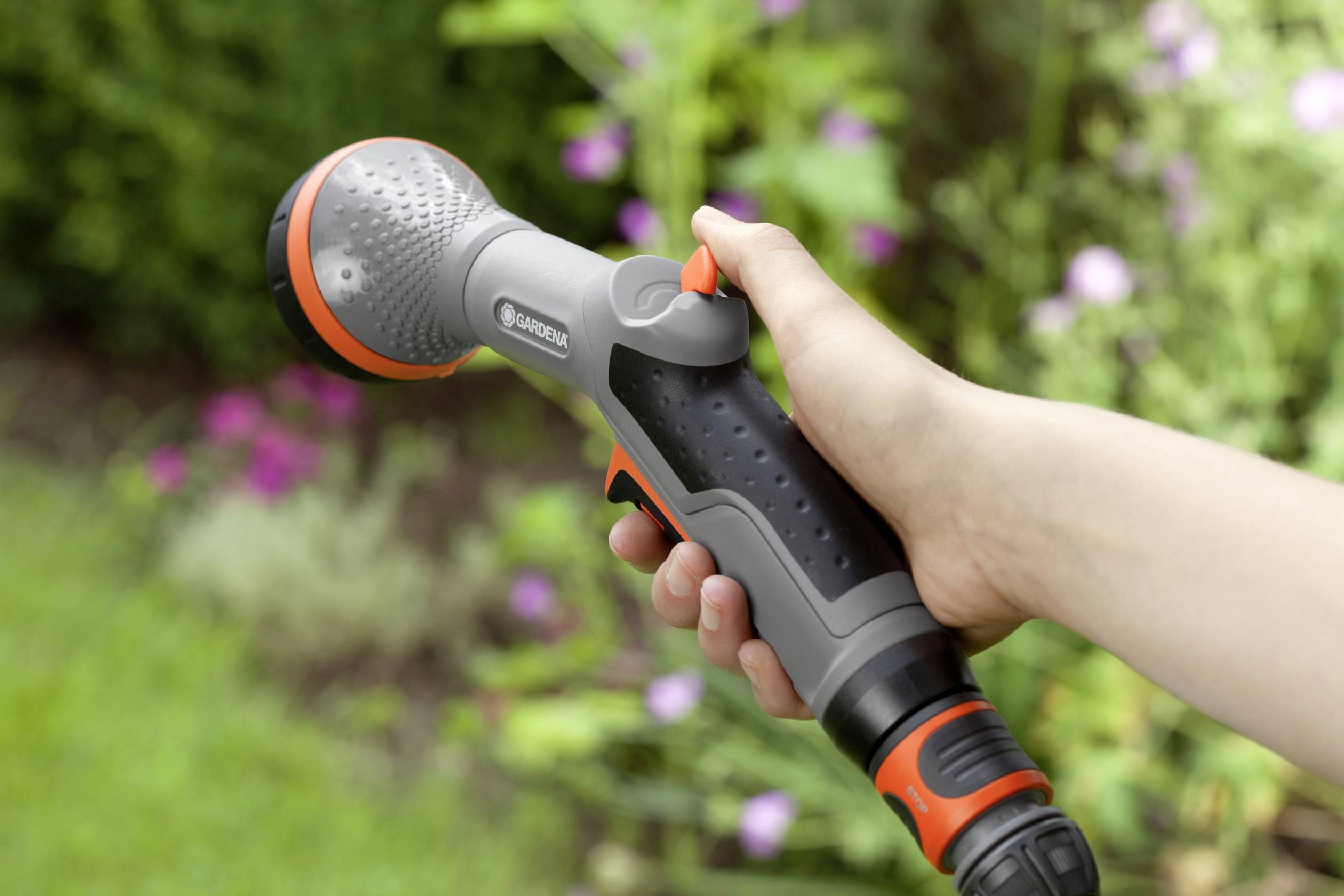 Dłoń trzyma szarą i pomarańczową pistolet do opryskiwania ogrodu z regulowaną dyszą, na rozmytym tle ogrodu.