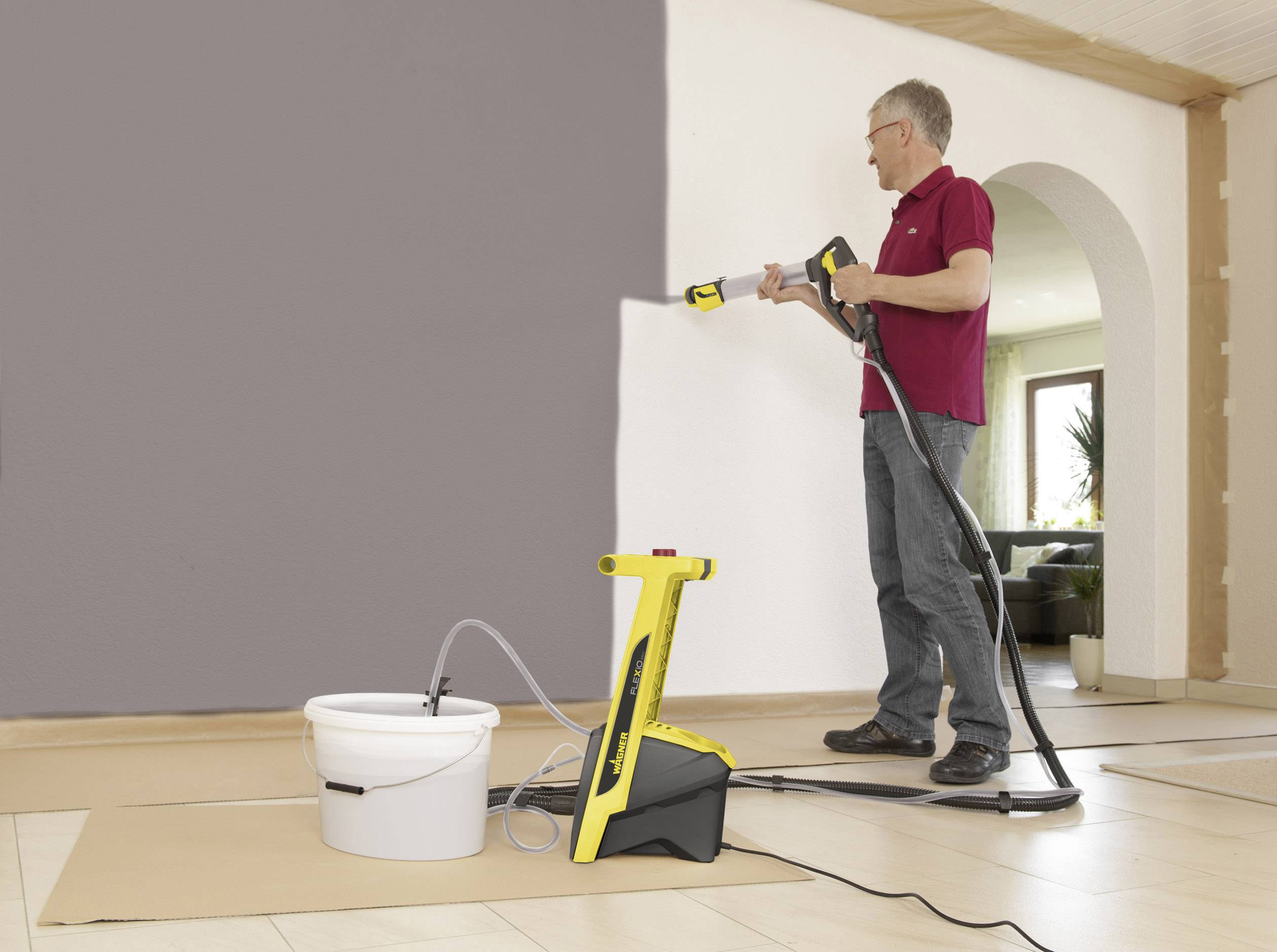 Mężczyzna maluje ścianę za pomocą żółtego pistoletu natryskowego. Przed nim stoi wiadro z farbą.