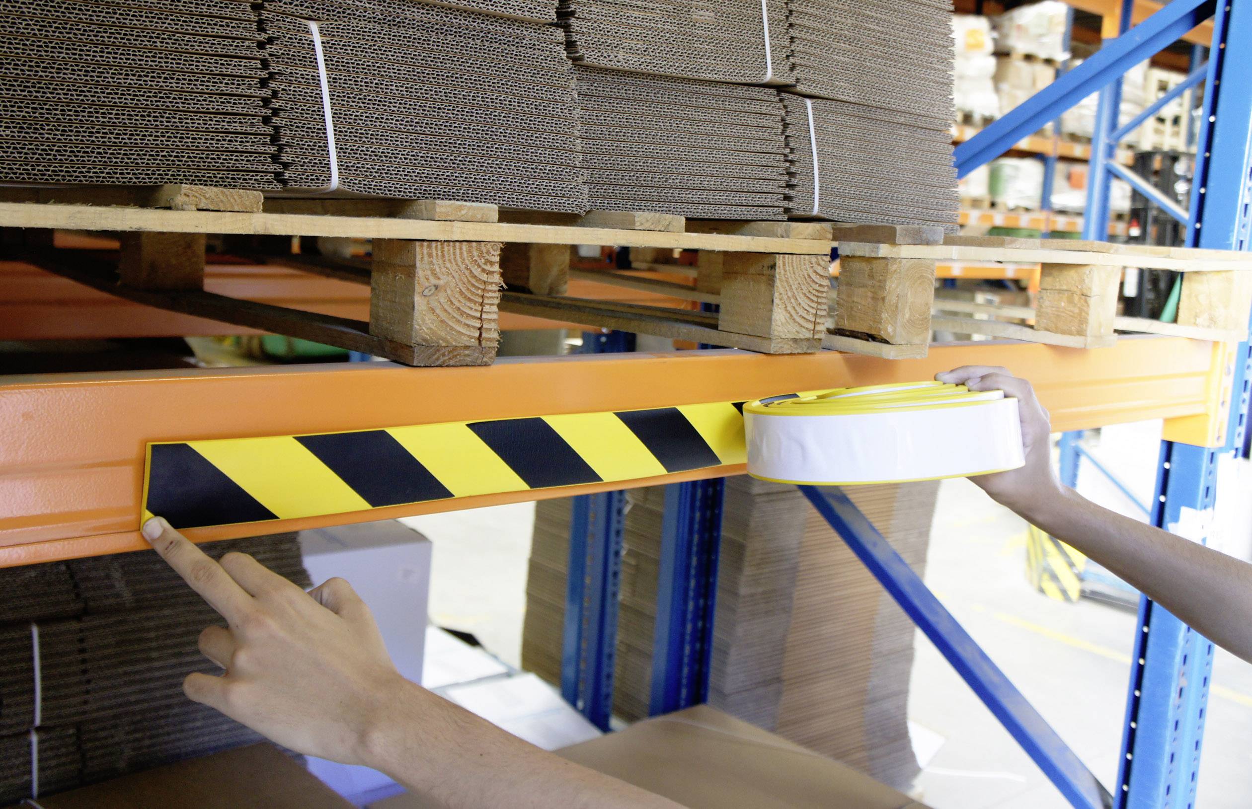 Osoba przymocowuje czarno-żółtą taśmę samoprzylepną do regału zawierającego kartony w magazynie.