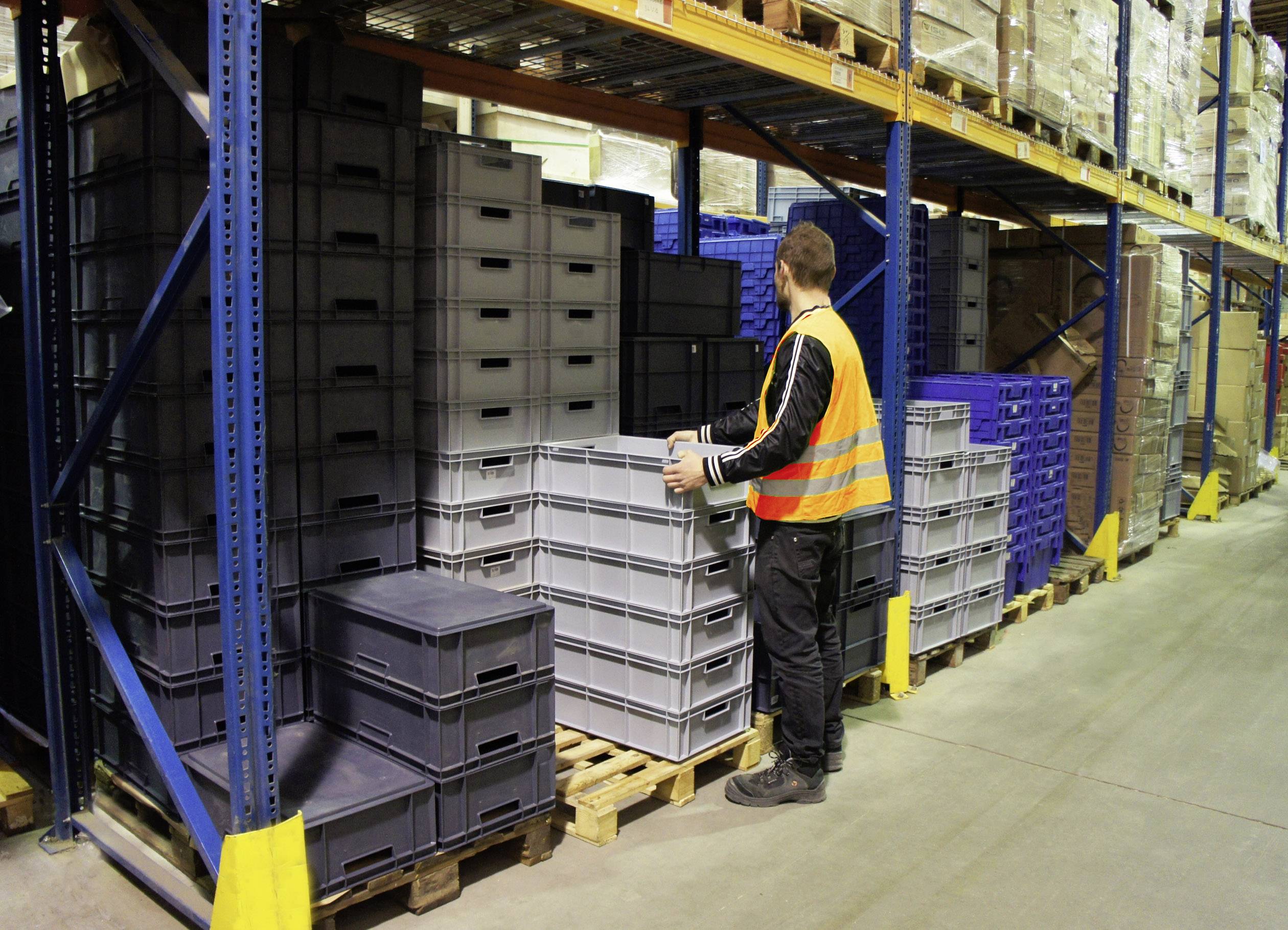 Robotnik w pomarańczowej kamizelce odblaskowej układa szare plastikowe skrzynki w magazynie z wysokimi regałami pełnymi kartonów.
