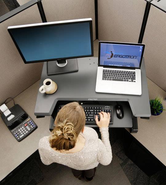 Osoba pracuje przy biurku do stania wyposażonym w dwa monitory, klawiaturę oraz kubek kawy. Otoczenie jest typowym stanowiskiem biurowym.