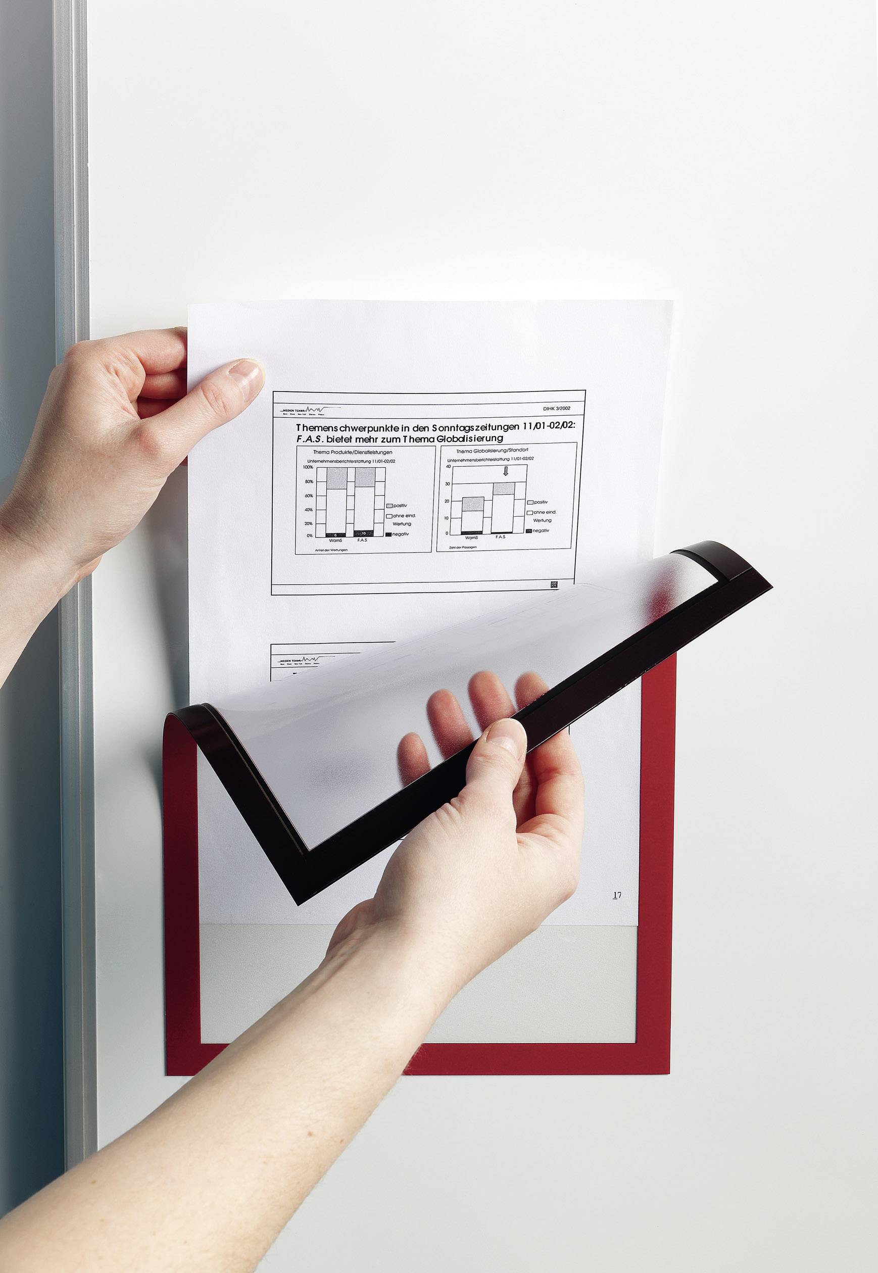 Dwie ręce trzymają kartkę papieru z diagramem, która jest umieszczana w przezroczystej osłonie z czerwoną ramką na ścianie.