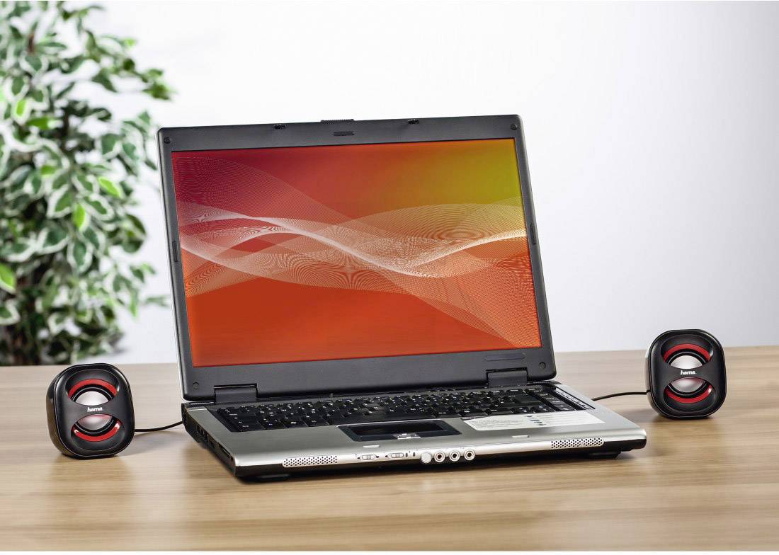 Laptop z otwartym ekranem wyświetla czerwoną, falistą grafikę tła. Dwa małe głośniki są umieszczone po lewej i prawej stronie. W tle widoczna jest część rośliny.