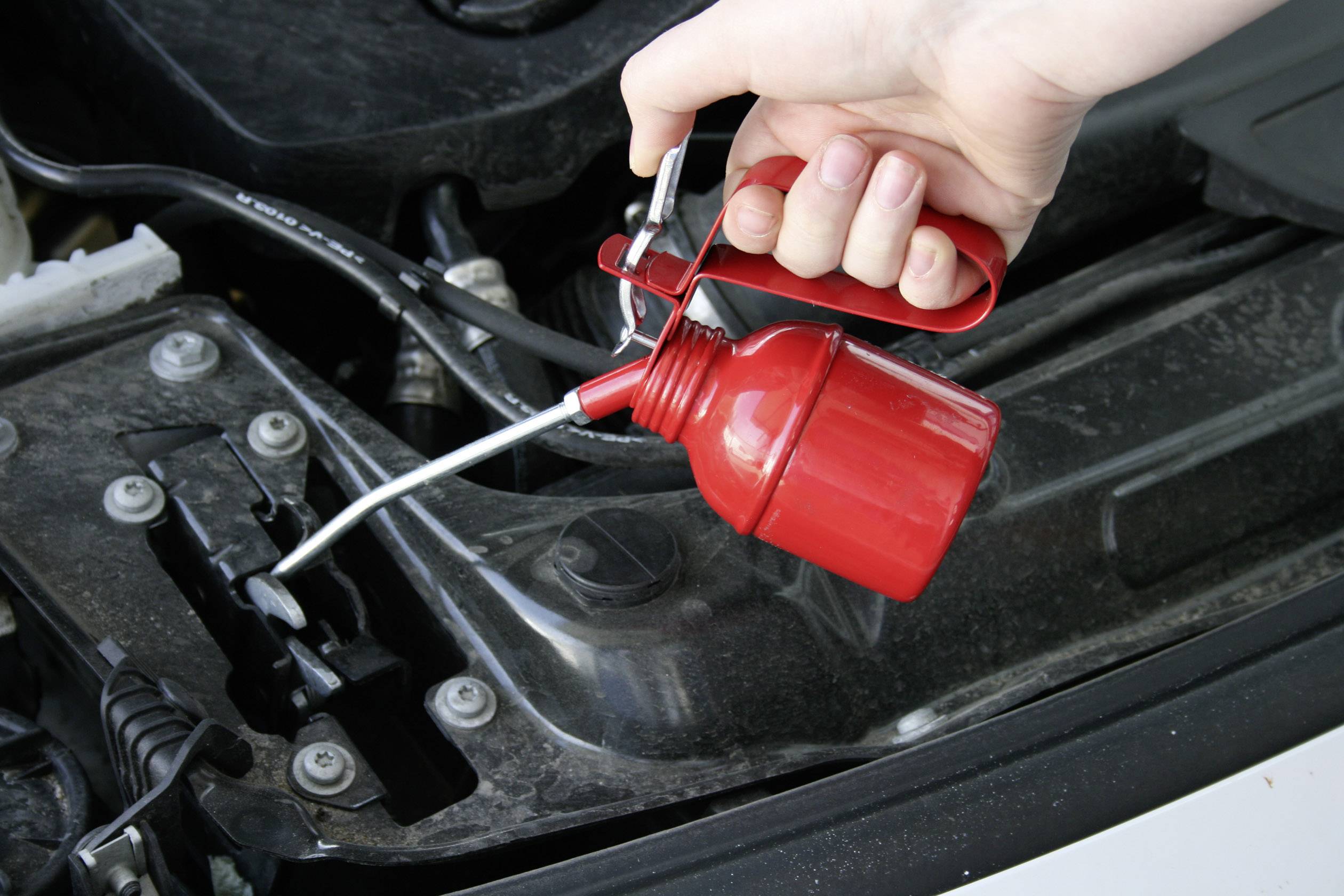 'Ktoś używa czerwonej puszki z olejem, aby nasmarować samochód przy masce.'