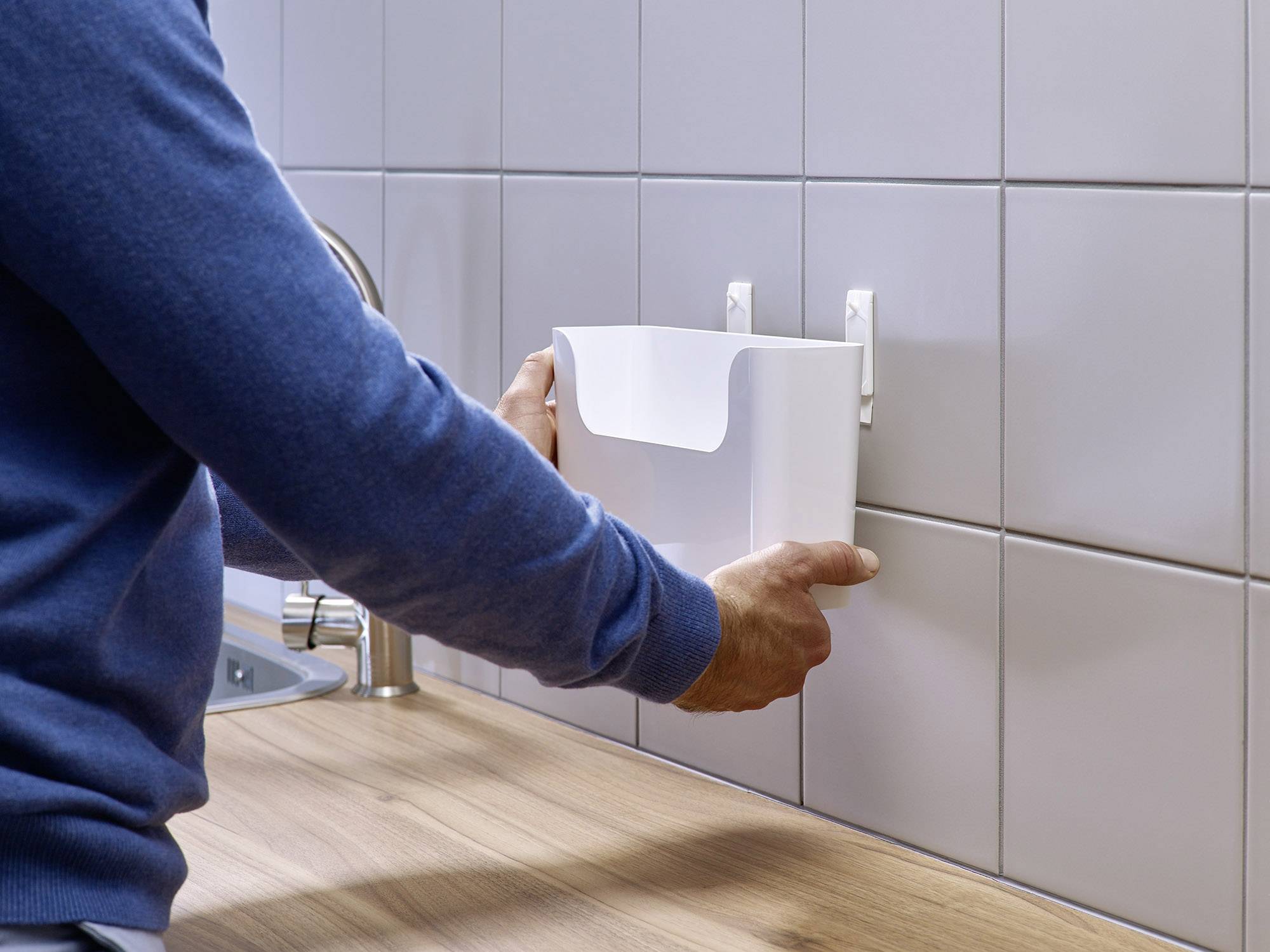 Osoba mocuje biały, wiszący kosz na śmieci na kafलowanej ścianie w pobliżu zlewu, który znajduje się na drewnianym blacie roboczym.