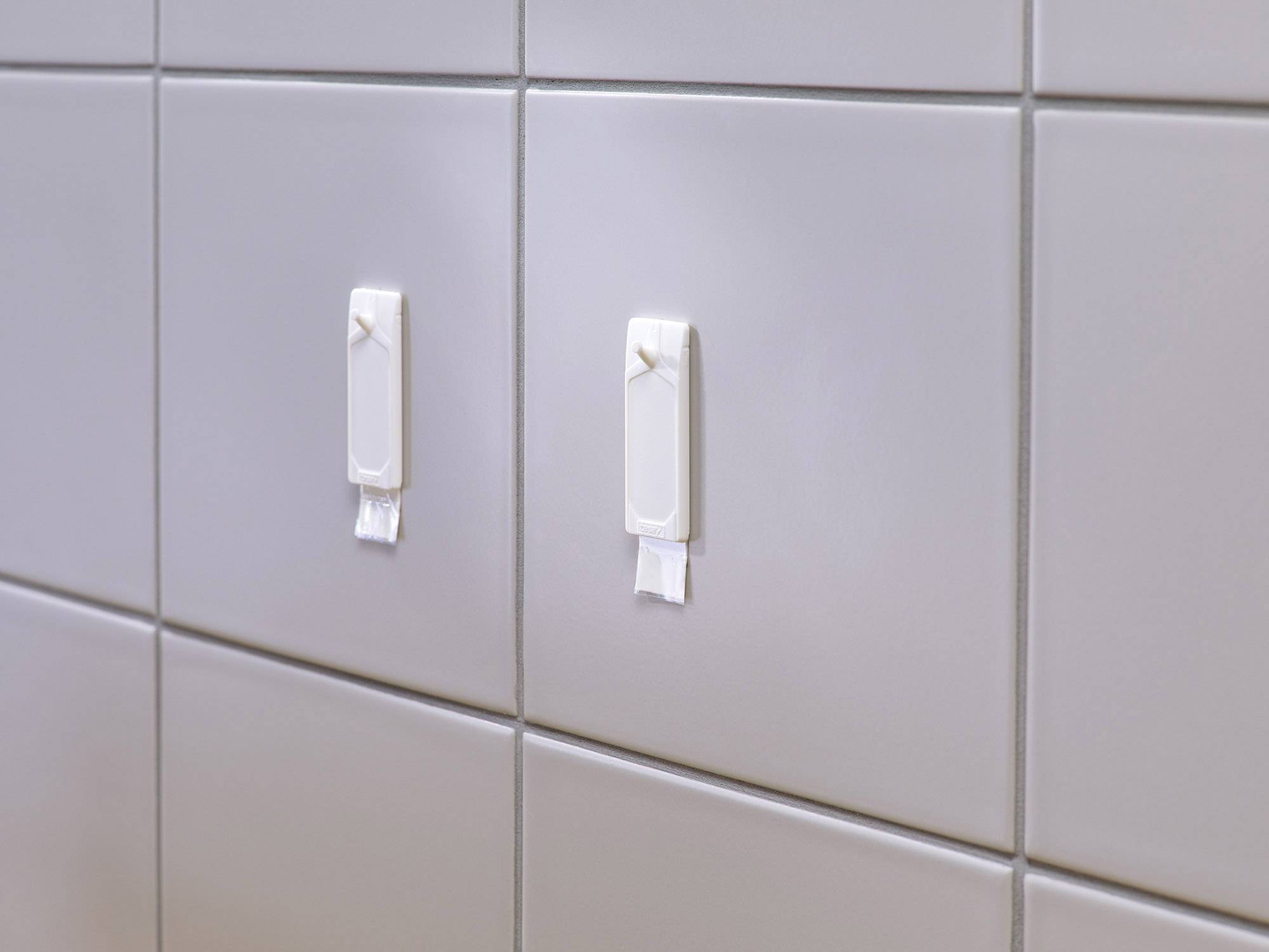 Dwa białe haczyki na jasnoszarych płytkach, przymocowane paskami klejącymi. Idealne do zawieszania lekkich przedmiotów w łazience.