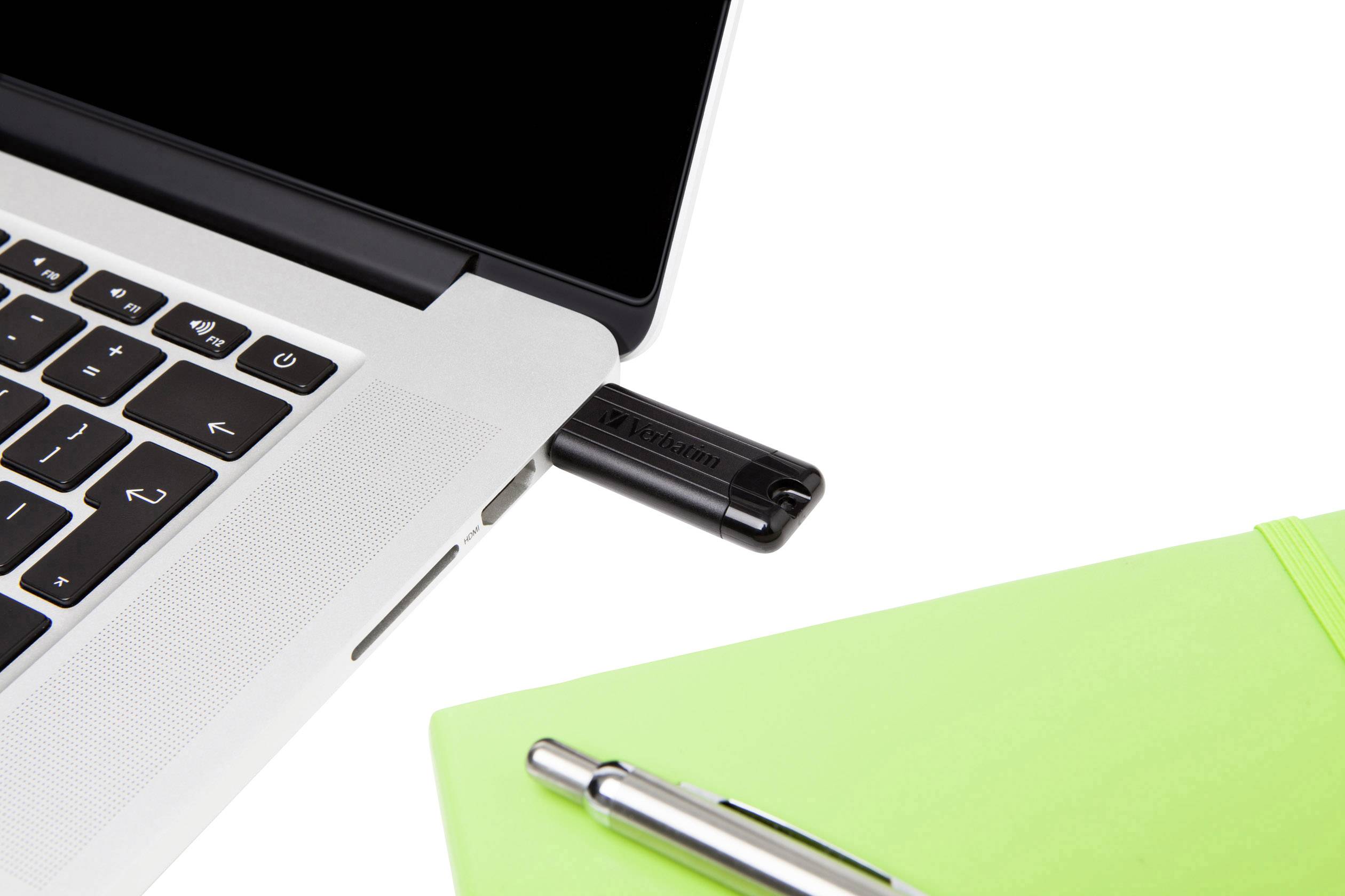 Laptop z podłączonym pendrivem USB obok zielonego notatnika i srebrnego długopisu na białym tle.