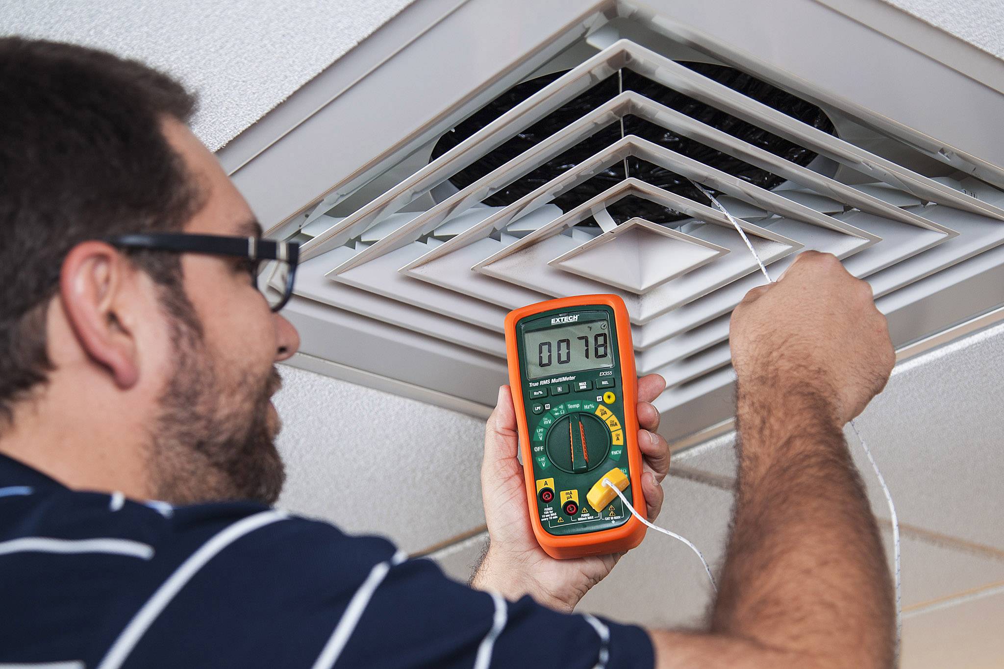 Osoba mierzy jakość powietrza w pobliżu kratki wentylacyjnej za pomocą urządzenia pomiarowego, które wskazuje wartość 0,178.