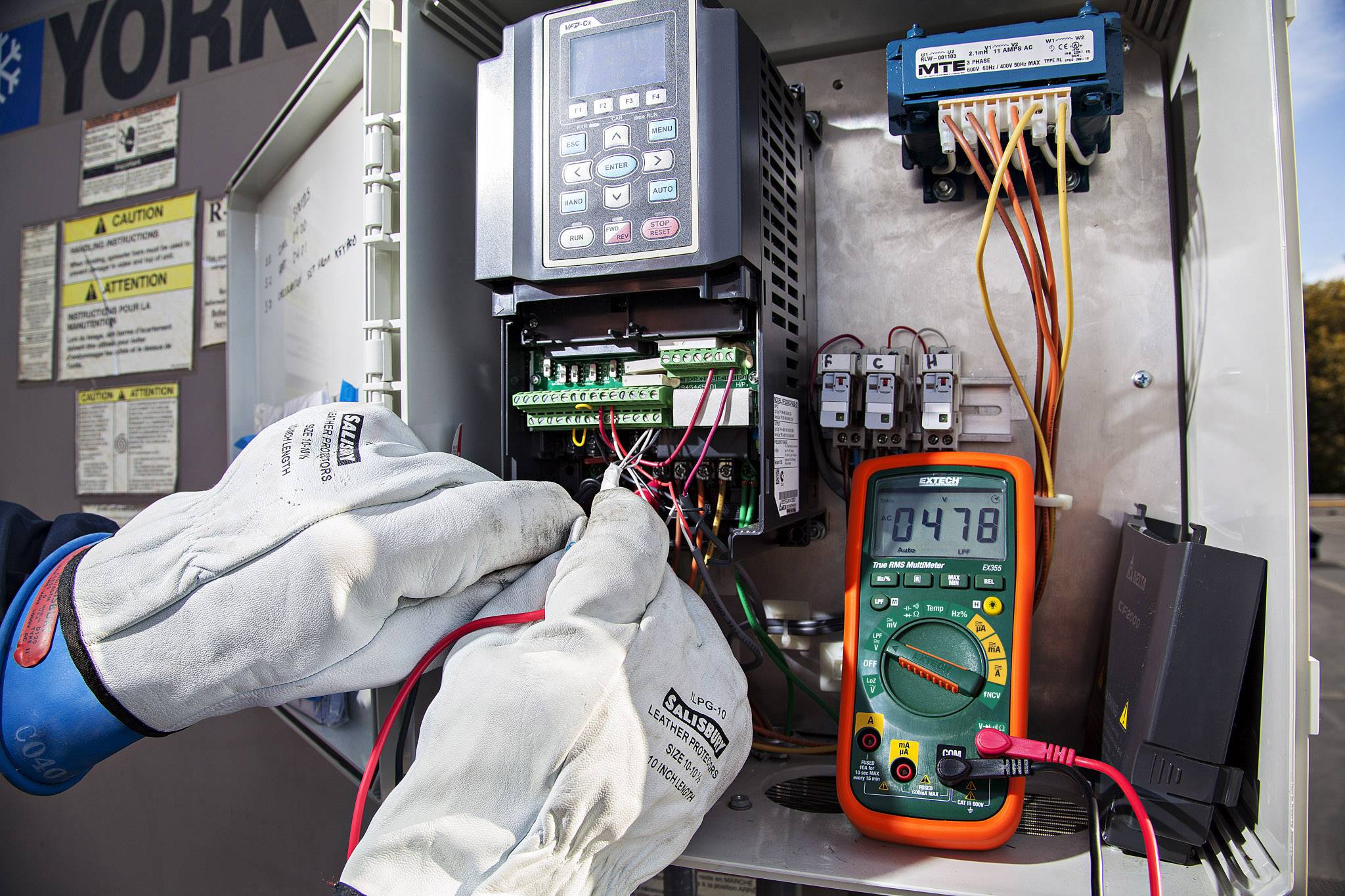 Elektryk mierzy napięcie przy otwartej szafce elektrycznej. Rękawice chronią przed porażeniem prądem. Multimetr wskazuje '0478'.