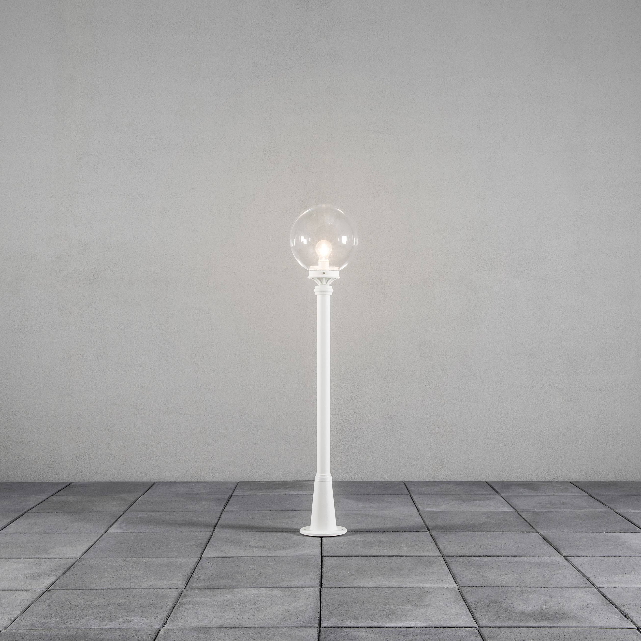 Biała lampa stojąca z kulistą żarówką stoi na szarej podłodze wyłożonej płytkami przed jasnoszarą ścianą.