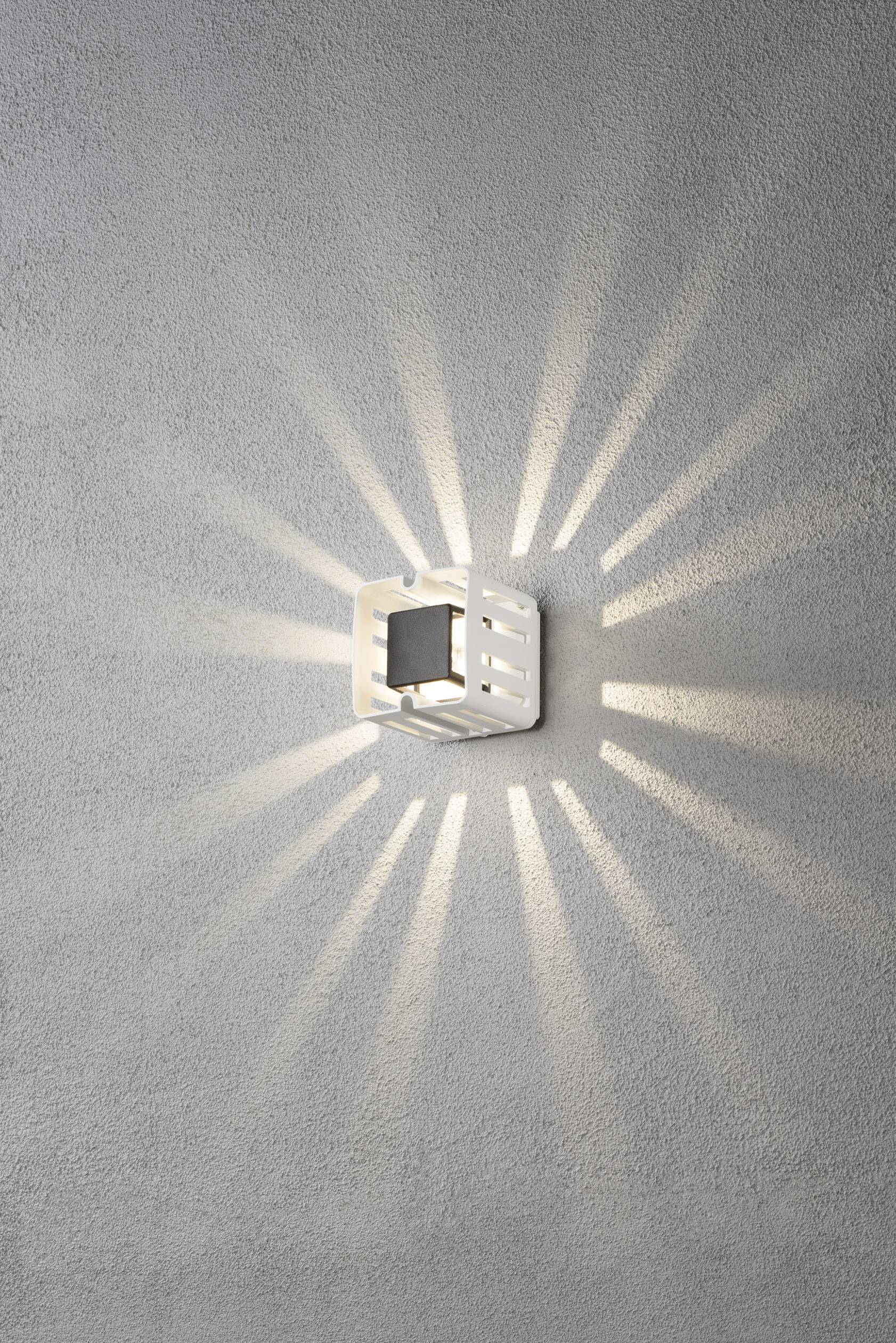 Oprawa ścienna rzuca symetryczne promienie światła, przypominające słońce. Lampa jest zamontowana w nowoczesnym stylu na szarej ścianie.