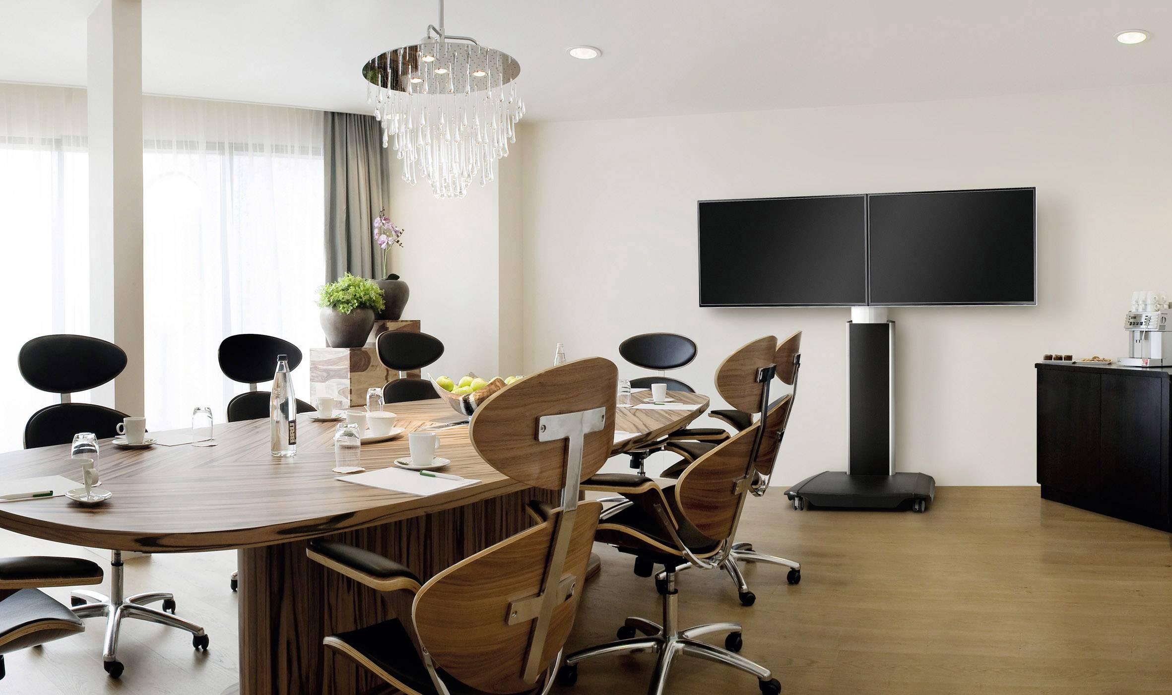 Sala konferencyjna z dużym drewnianym stołem, nowoczesnymi krzesłami, dwoma dużymi ekranami na jednym stojaku oraz dekoracją roślinną.