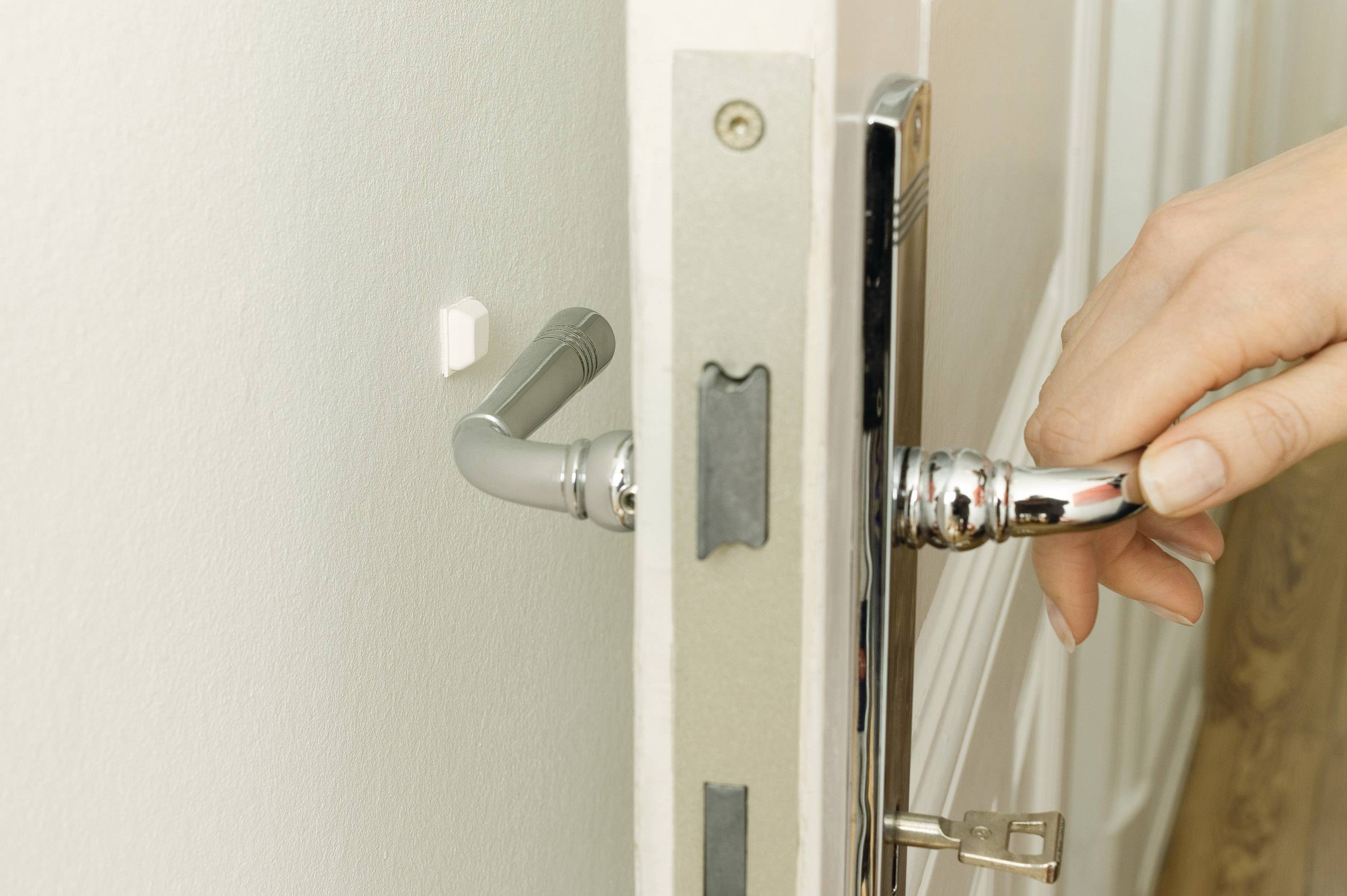 Ręka otwiera białe drzwi pokojowe z srebrną klamką. Klucz jest włożony do zamka.