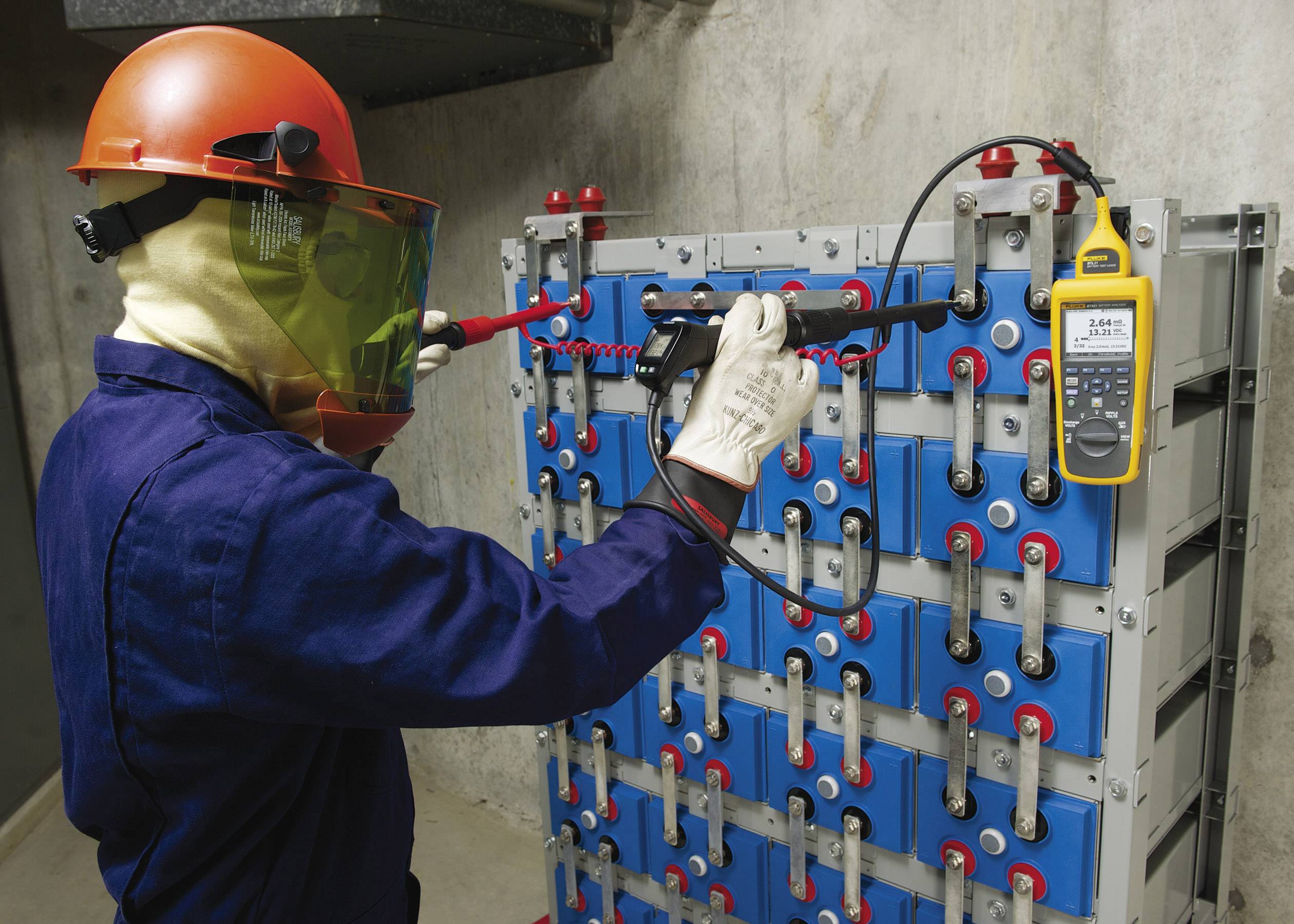 Osoba w odzieży ochronnej sprawdza napięcie przy pomocy urządzenia na serii niebieskich baterii w betonowym pomieszczeniu.