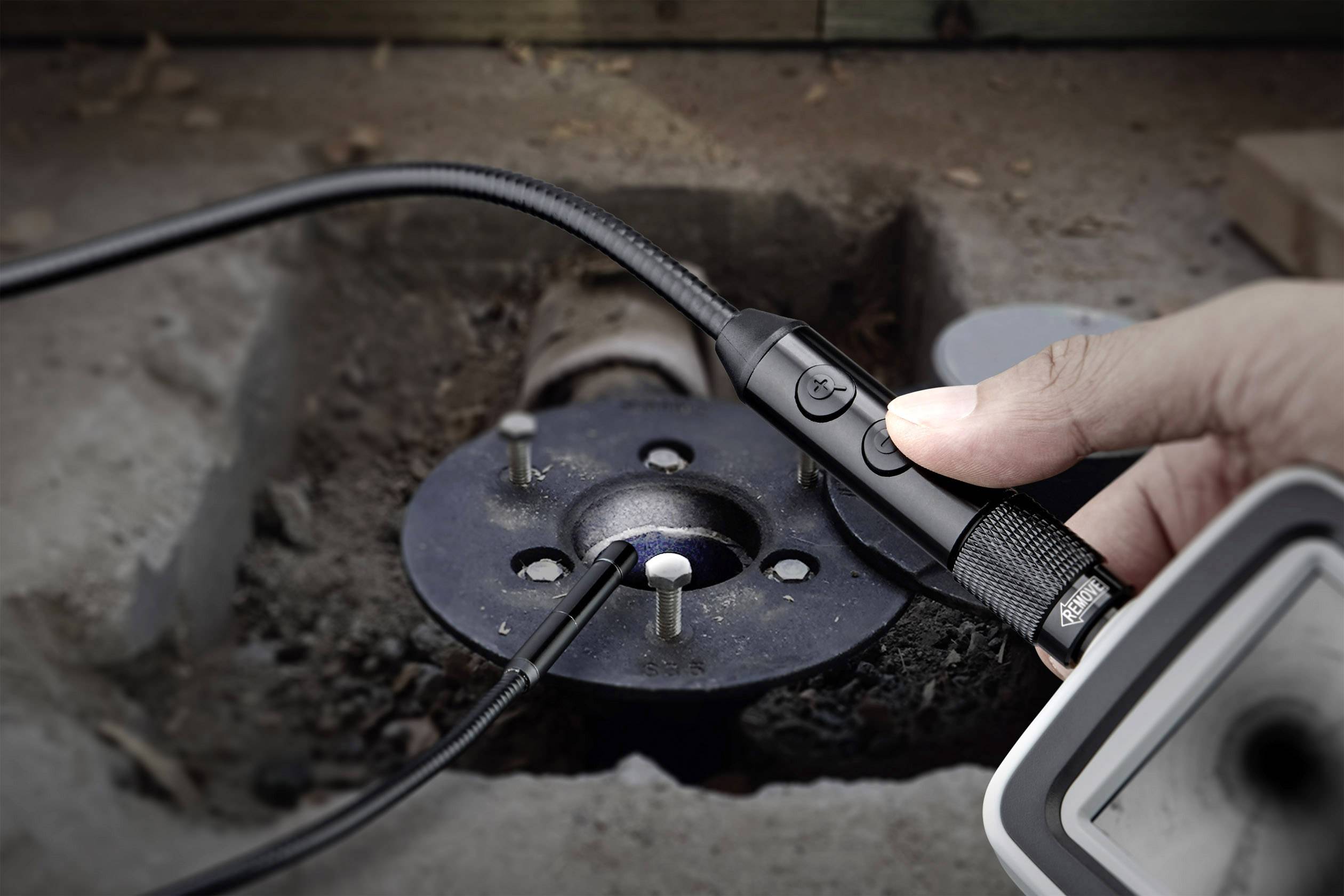 Dłoń trzyma kamerę inspekcyjną nad ciemnym otworem w betonowej podłodze. Kamera jest przymocowana do elastycznego kabla, umożliwiającego badanie trudno dostępnych miejsc.