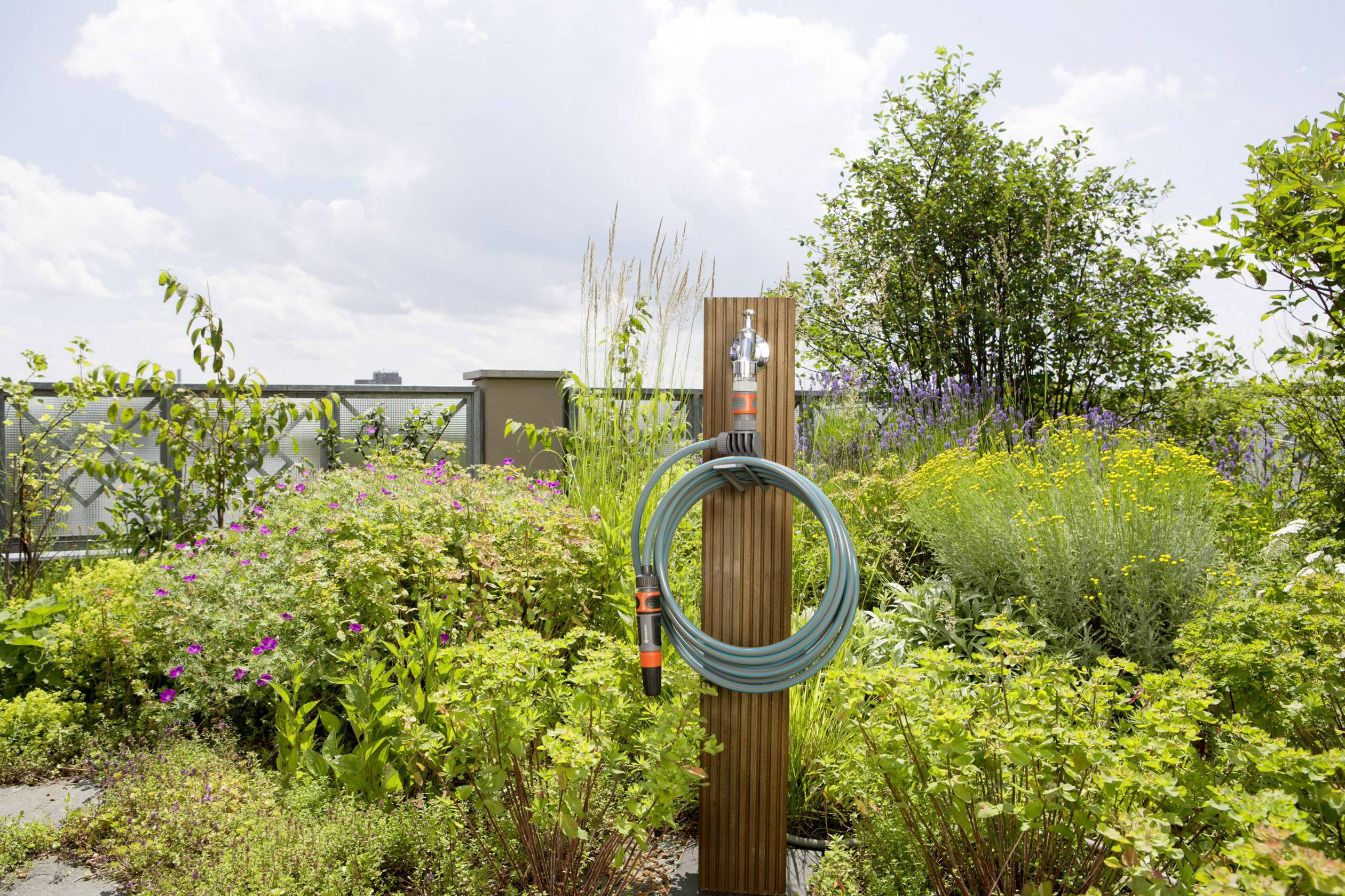 Wąż ogrodowy jest owinięty wokół drewnianego słupa, otoczony bujną zielenią i kolorowymi kwiatami. W tle widać pochmurne niebo.