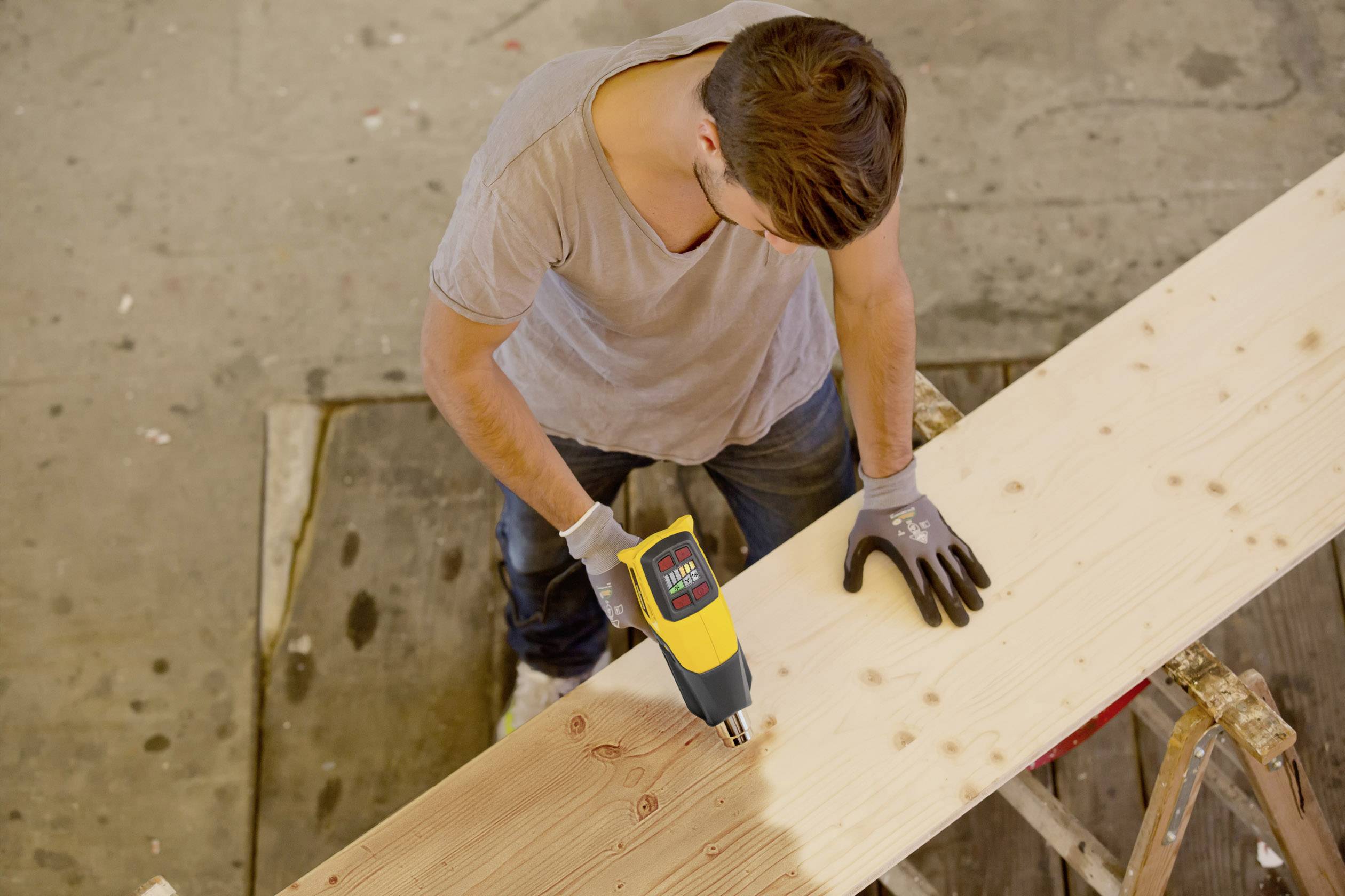 Mężczyzna szlifuje deskę drewnianą elektronarzędziem w warsztacie. Nosi rękawice ochronne.