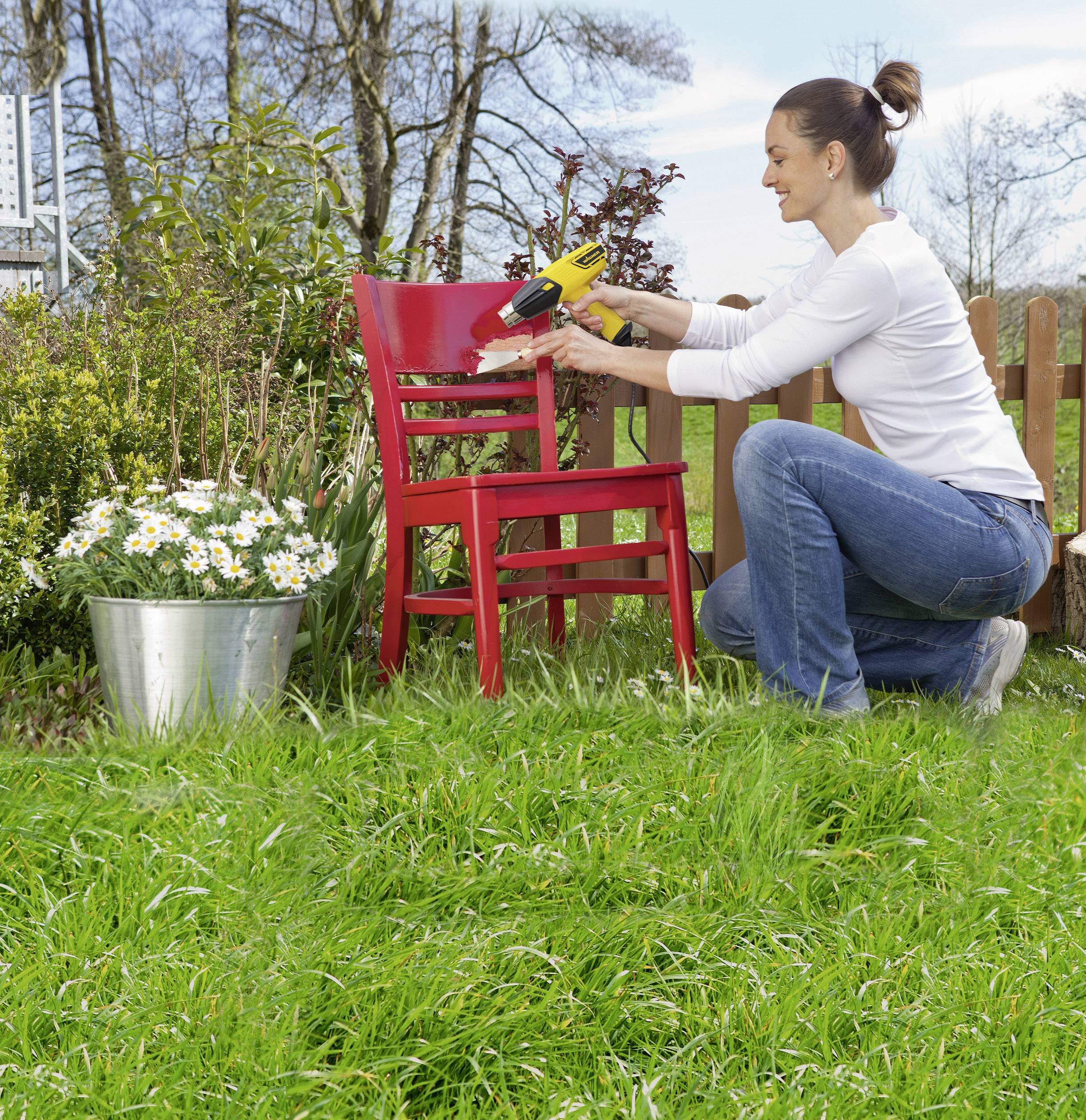 Kobieta usuwa farbę z czerwonego krzesła za pomocą elektronarzędzia w ogrodzie. W tle widoczne są kwiaty i drewniany płot.