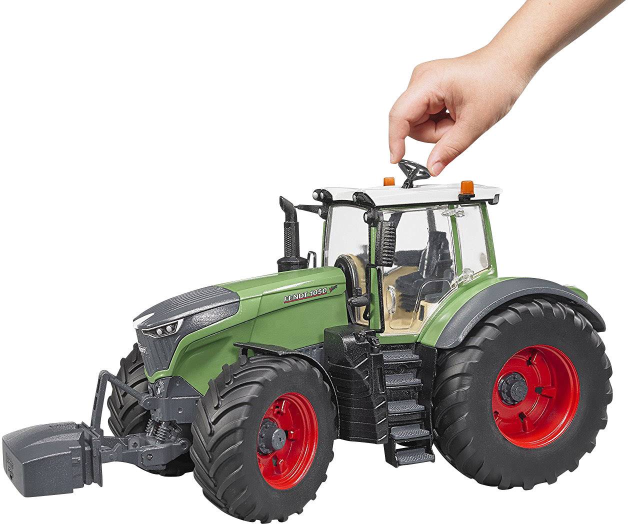Miniaturowy traktor w zielonym kolorze nadwozia i czerwonych kołach. Ręka obraca kierownicą na górze traktora.