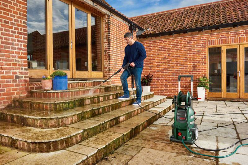 Osoba czyści wielostopniowe kamienne schody przed czerwonym domem z cegły za pomocą myjki ciśnieniowej.