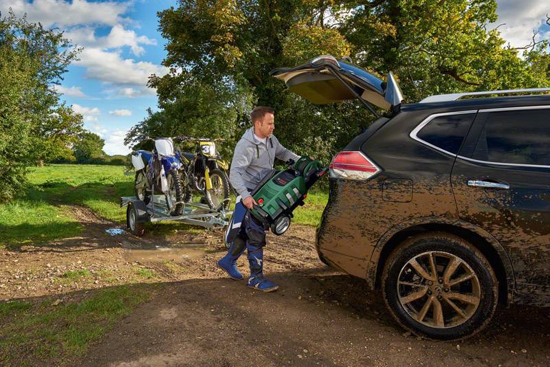 Mężczyzna ładuje plecak do bagażnika SUV-a. W tle widać dwa motocykle na przyczepie w wiejskiej scenerii.