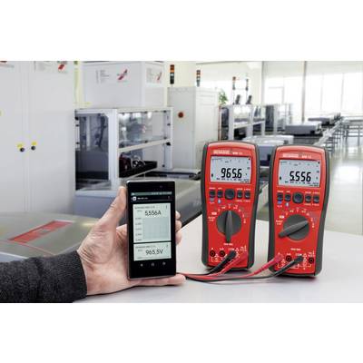 Jedna ręka trzyma smartfon z wyświetlanymi cyframi, obok stoją dwa cyfrowe multimetry na stole w środowisku laboratoryjnym.