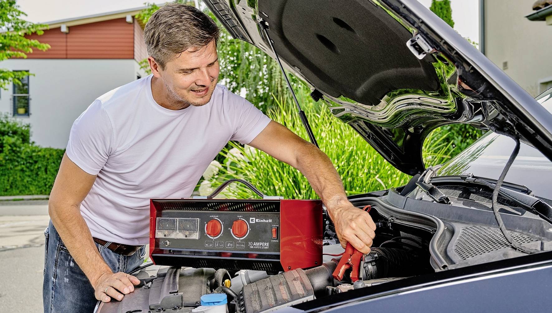 Mężczyzna użycza rozruchu samochodowi na zewnątrz przy pomocy przewodów rozruchowych. Maska silnika jest otwarta; mężczyzna ma na sobie białą koszulkę.