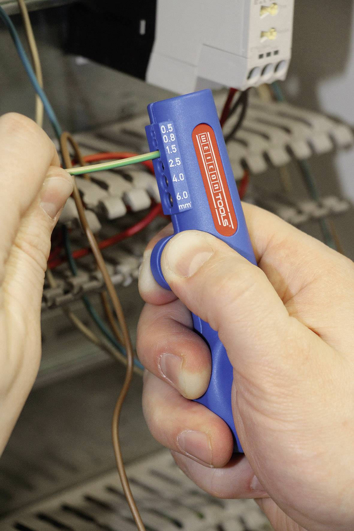 Osoba trzyma niebieskie szczypce do zdejmowania izolacji, aby odsłonić przewód elektryczny w szafce rozdzielczej.