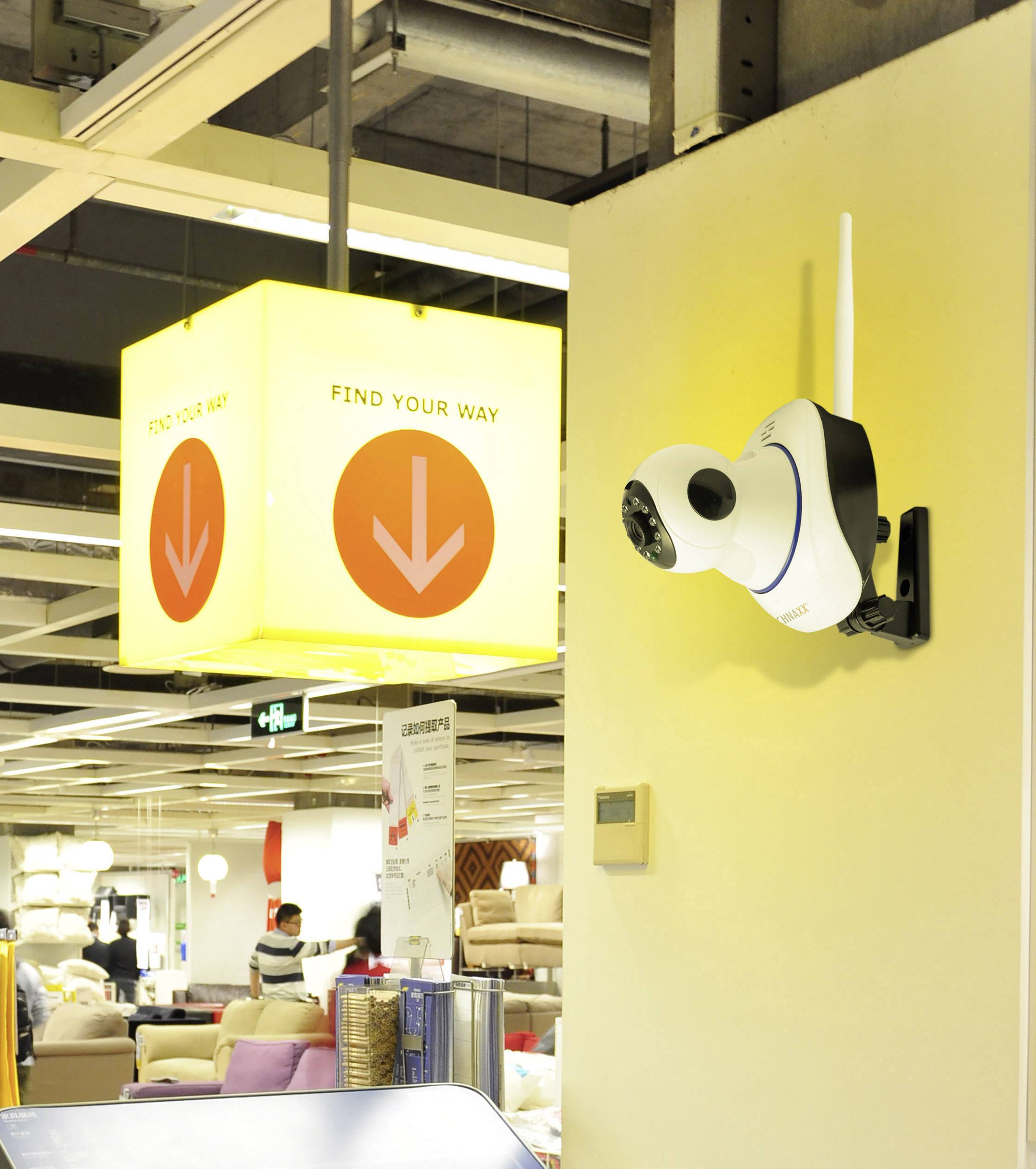 Kamera monitorująca zamontowana na ścianie wewnętrznej, nadzoruje obszar w sklepie meblowym. Tabliczka z napisem 'FIND YOUR WAY' i strzałkami zwisa z sufitu.