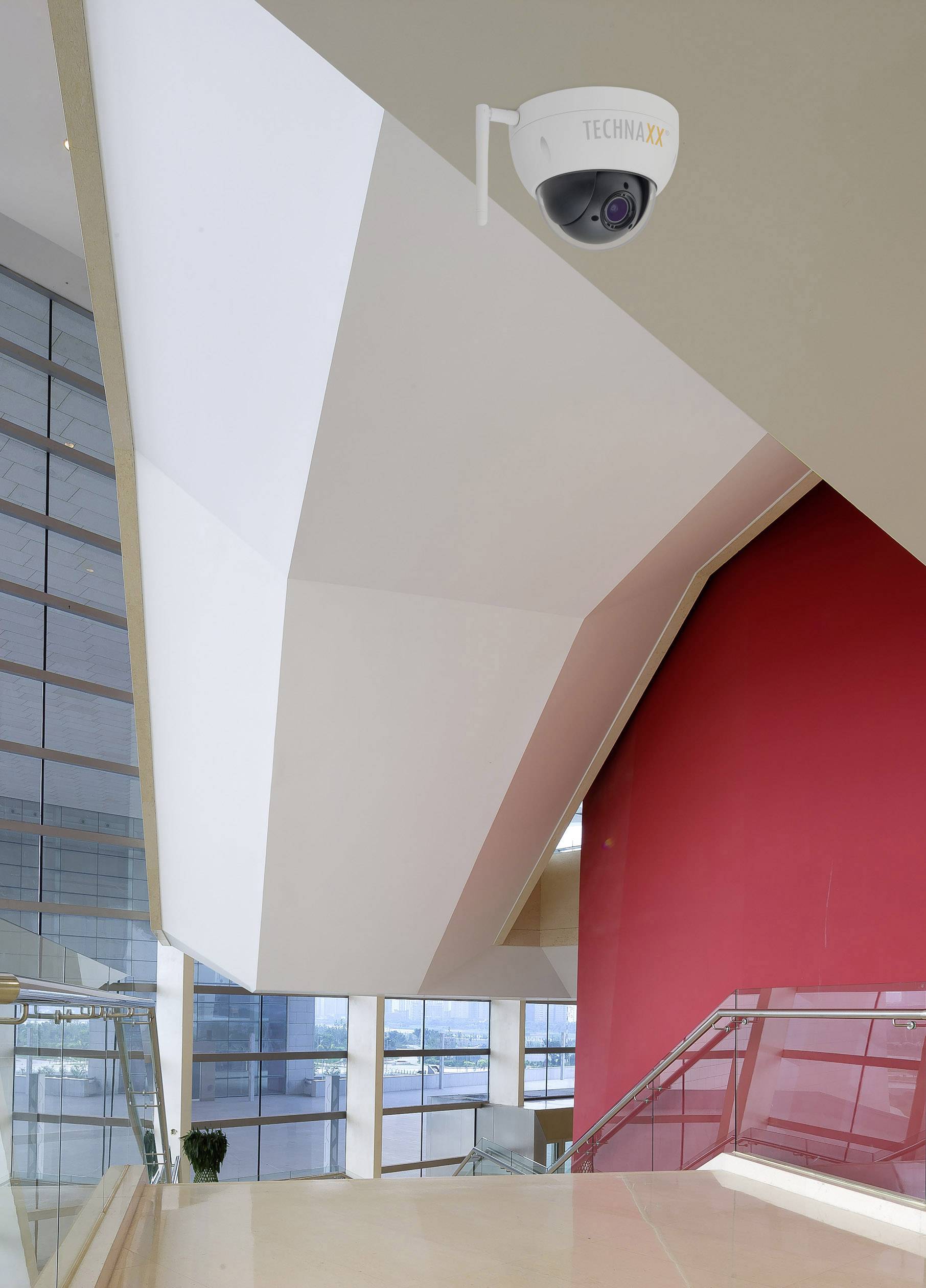 Widok wnętrza nowoczesnego budynku z kamerą monitoringu na suficie, czerwonymi ścianami i dużymi oknami z widokiem na zewnątrz.