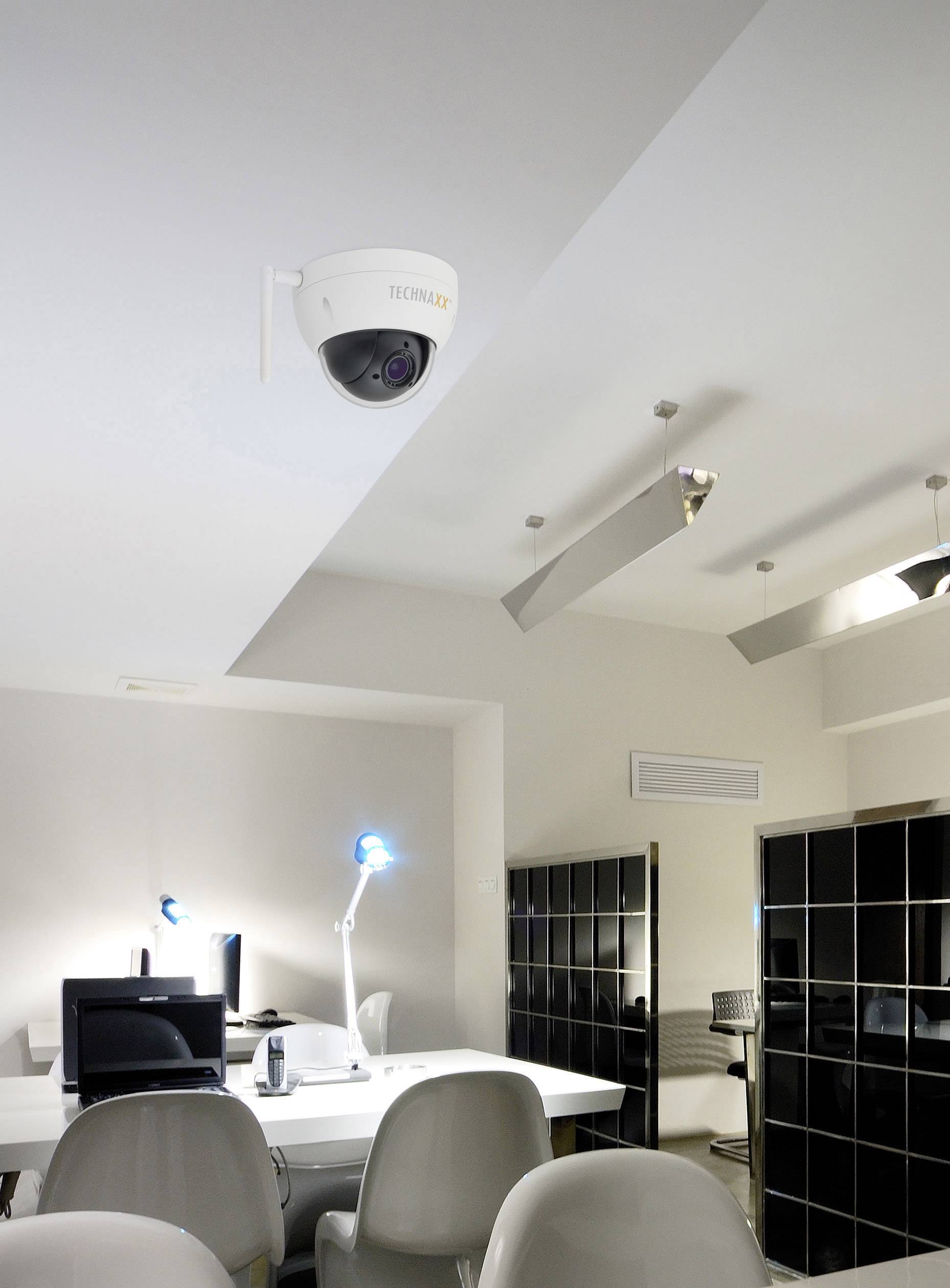 Biuro z białymi stołami i szarymi krzesłami. Na suficie zamontowana jest kamera monitoringu. W tle stoją komputery z niebieską lampką biurkową.