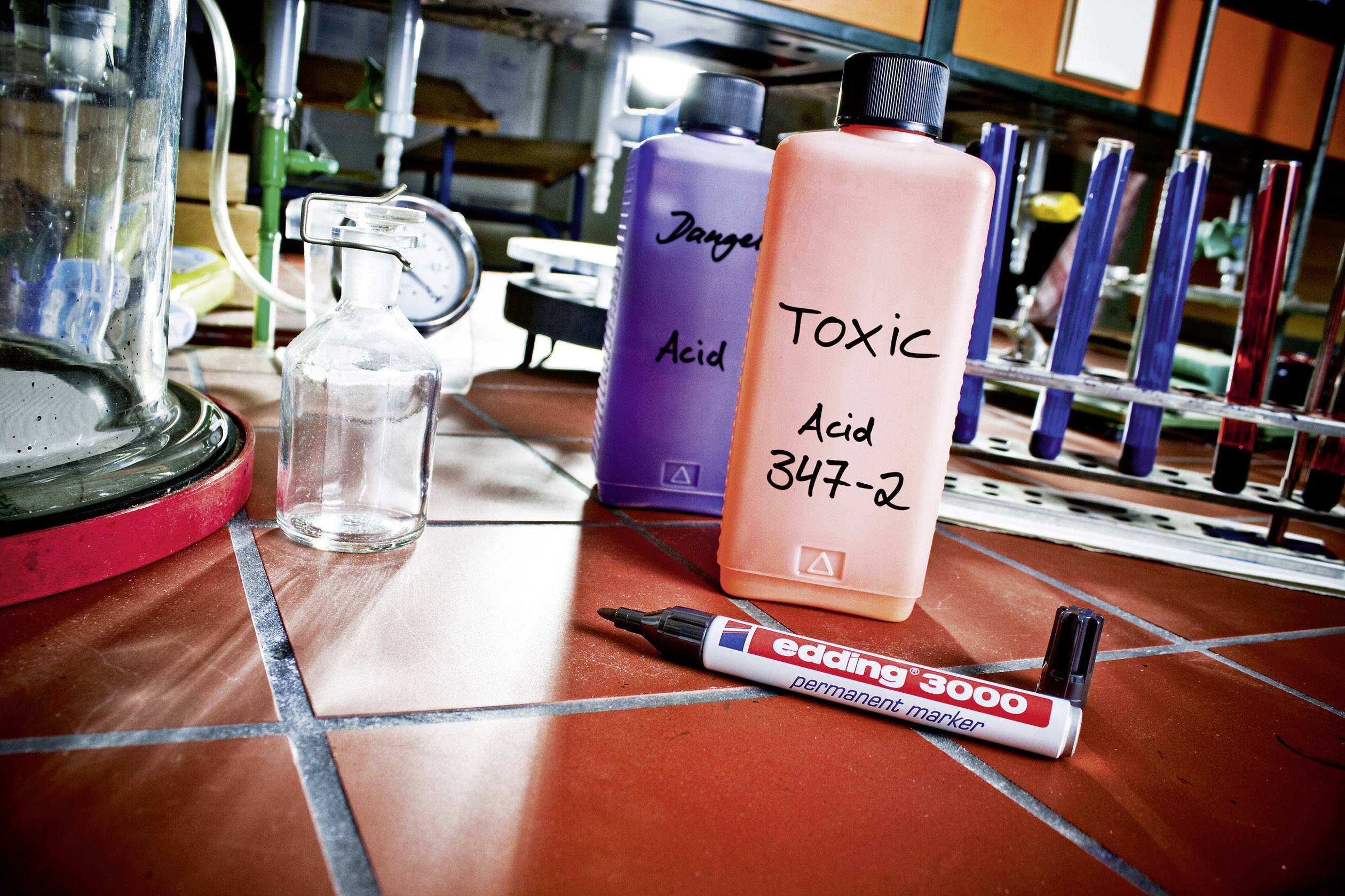 Dwie butelki z napisami 'Danger Acid' i 'Toxic Acid 347-2' stoją na podłodze wyłożonej płytkami w laboratorium, obok markera.
