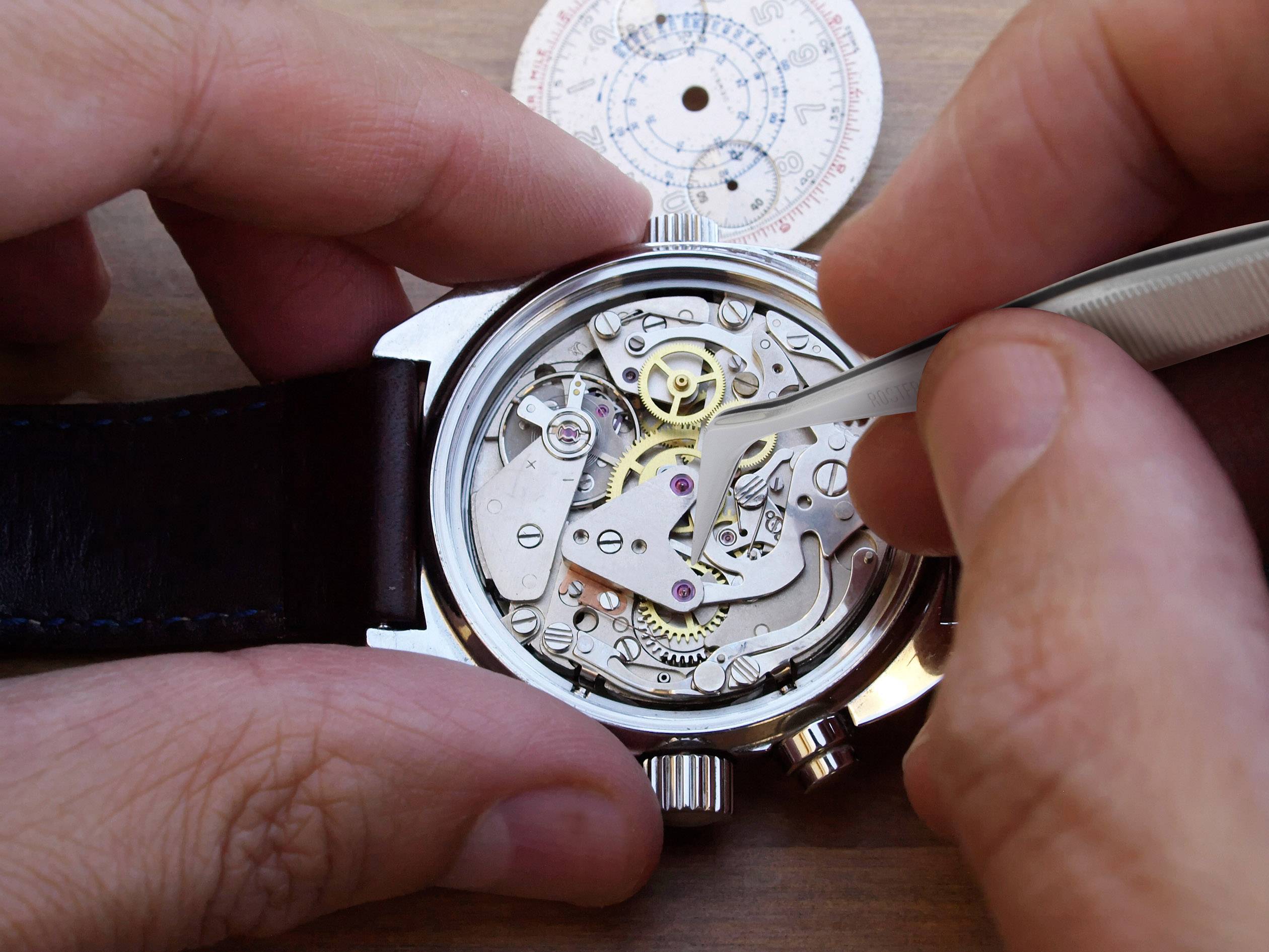 Osoba naprawia mechanizm zegarka za pomocą pincety. Widoczne są kółka zębate i drobne detale.