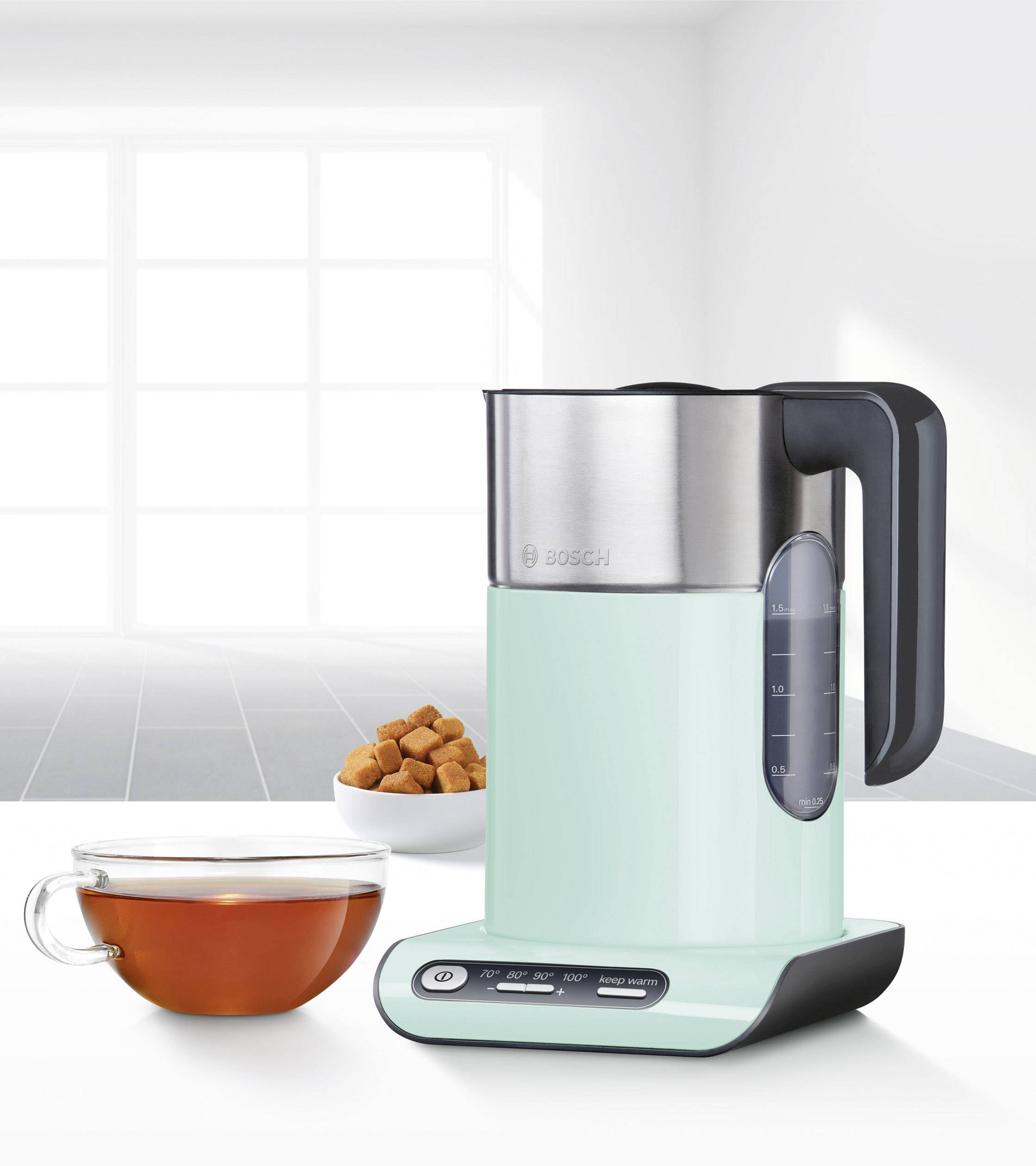 Czajnik elektryczny stojący na stole przed oknem. Obok znajduje się miska z kostkami cukru i filiżanka herbaty.