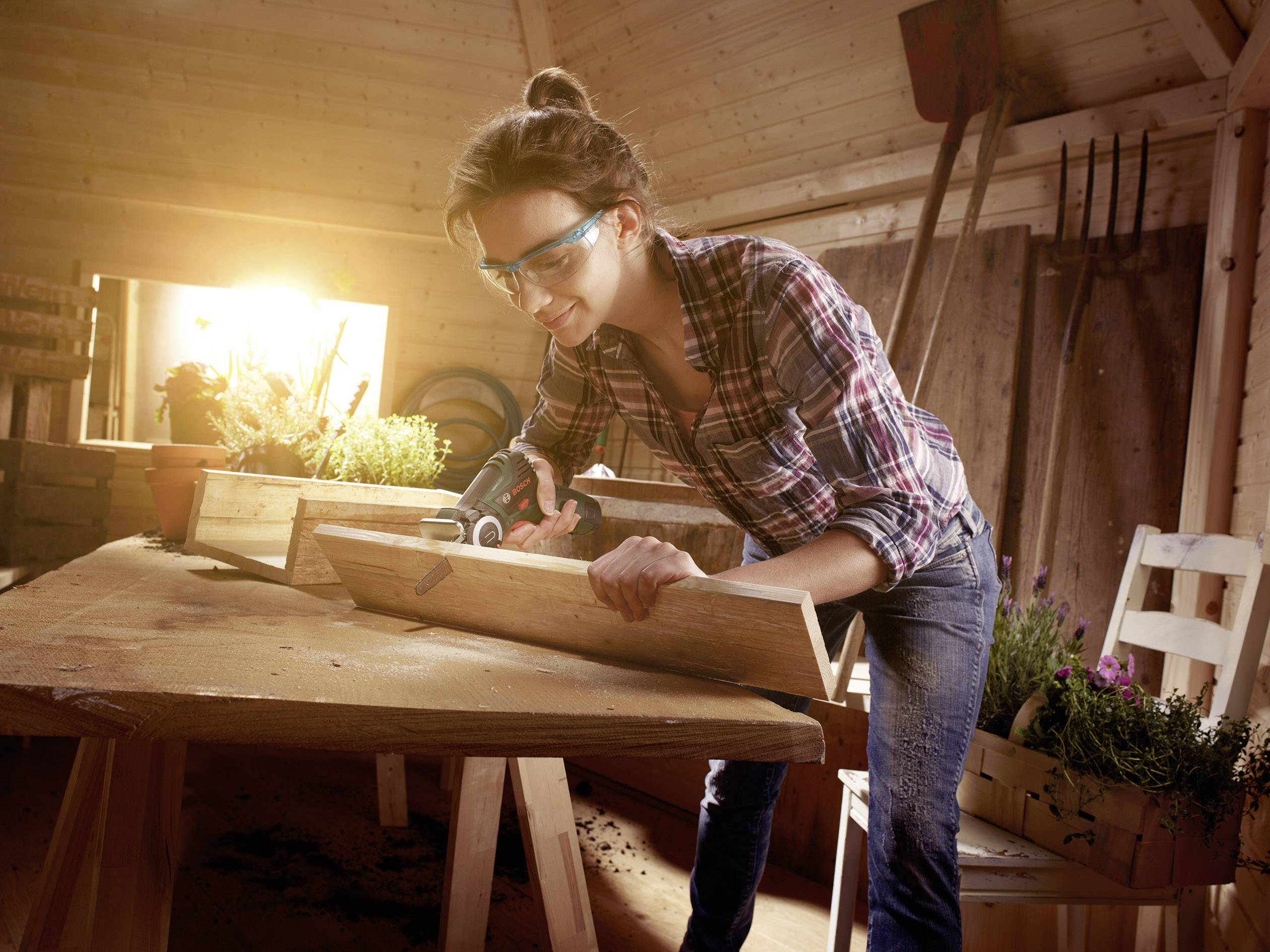 Osoba pracuje w warsztacie stolarskim w ochronnych okularach, piłuje deskę na stole. Przez okno pada słoneczne światło.